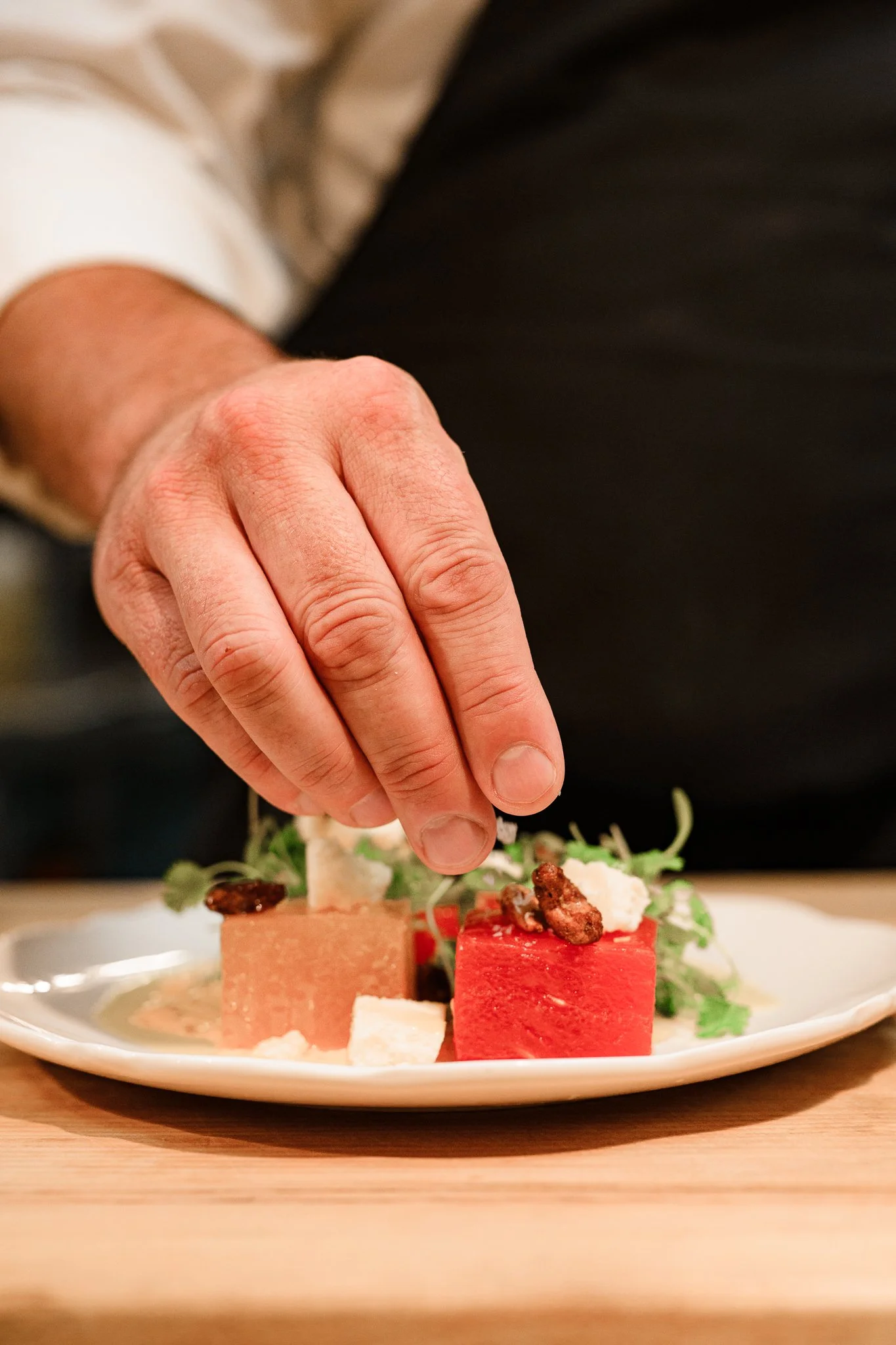 Close-up of a hand placing a topping on a plate of assorted food items, including pieces of watermelon and cheese, with some greens in the background.