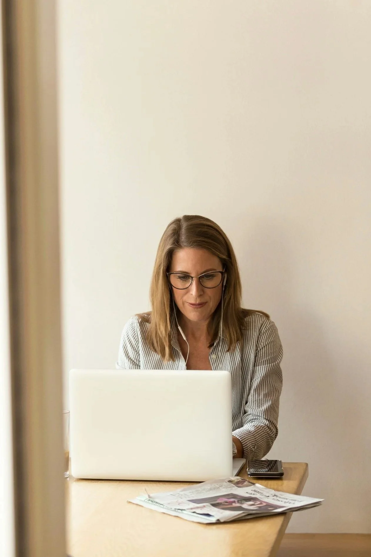 A woman with glasses and earphones wearing a striped shirt is working on a laptop at a desk. Newspapers and a phone are on the desk. The photo is taken through a partially opened door with a beige wall in the background.