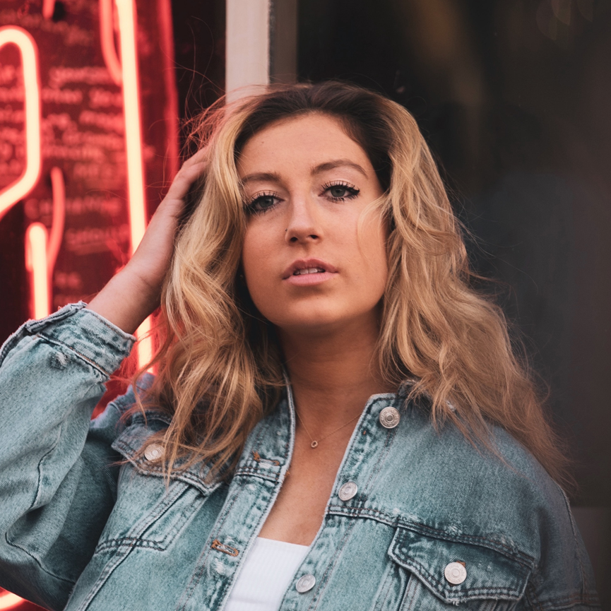 A young woman with wavy blonde hair wearing a denim jacket, standing in front of a neon sign.