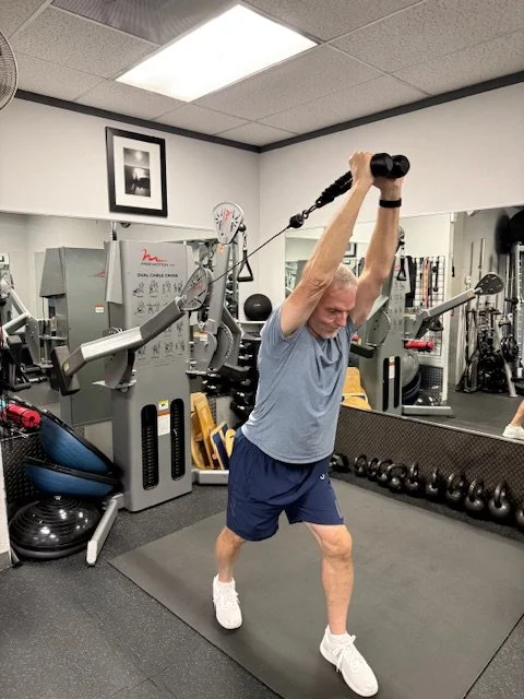 An elderly man lifting a weight with a handle over his head in a gym. He is standing on a black exercise mat, surrounded by gym equipment.