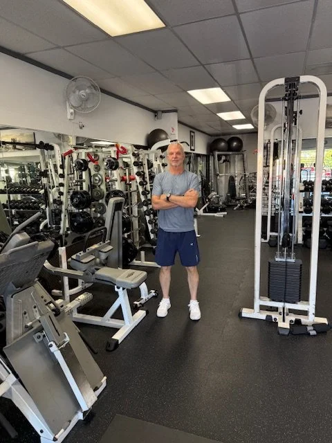 A man standing with arms crossed inside a gym with various weightlifting equipment including dumbbells, machines, and a cable machine.