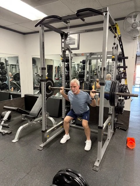 Man in gray shirt and shorts doing squats with a barbell on a power rack in a gym.