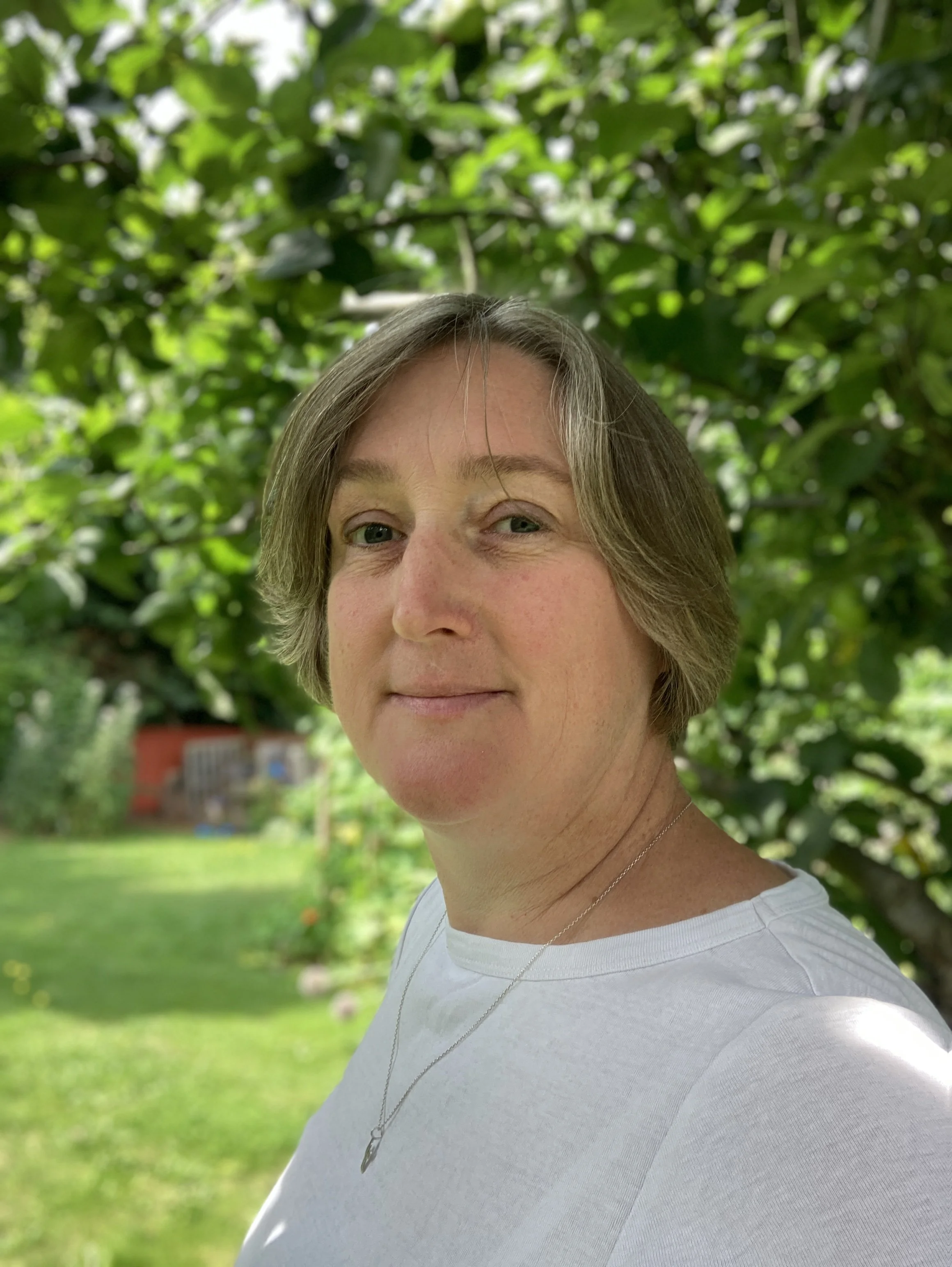 A woman with short brown hair standing outdoors in front of green foliage, wearing a white shirt and a necklace, with a blurred background of a garden.