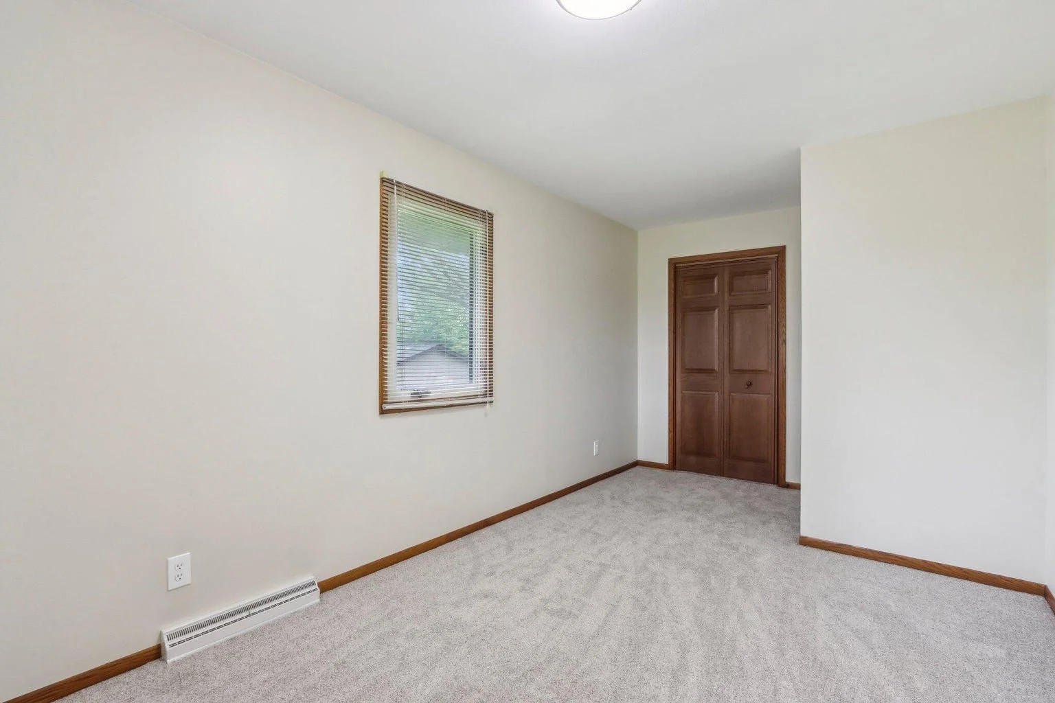 Empty room with beige carpet, cream walls, a window with blinds, and a wooden closet door.