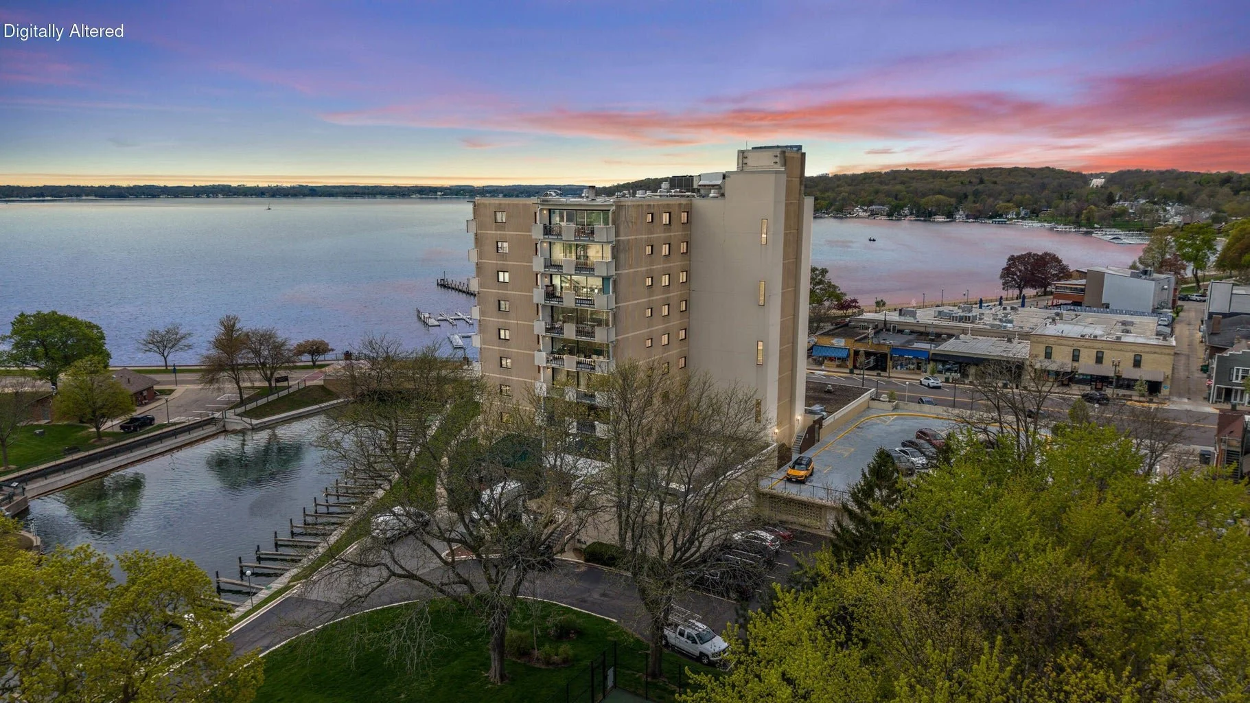 A high-rise apartment building overlooks a water lake with docks and boats, surrounded by trees, parking lots, and small shops on a scenic riverfront during sunset with colorful sky.