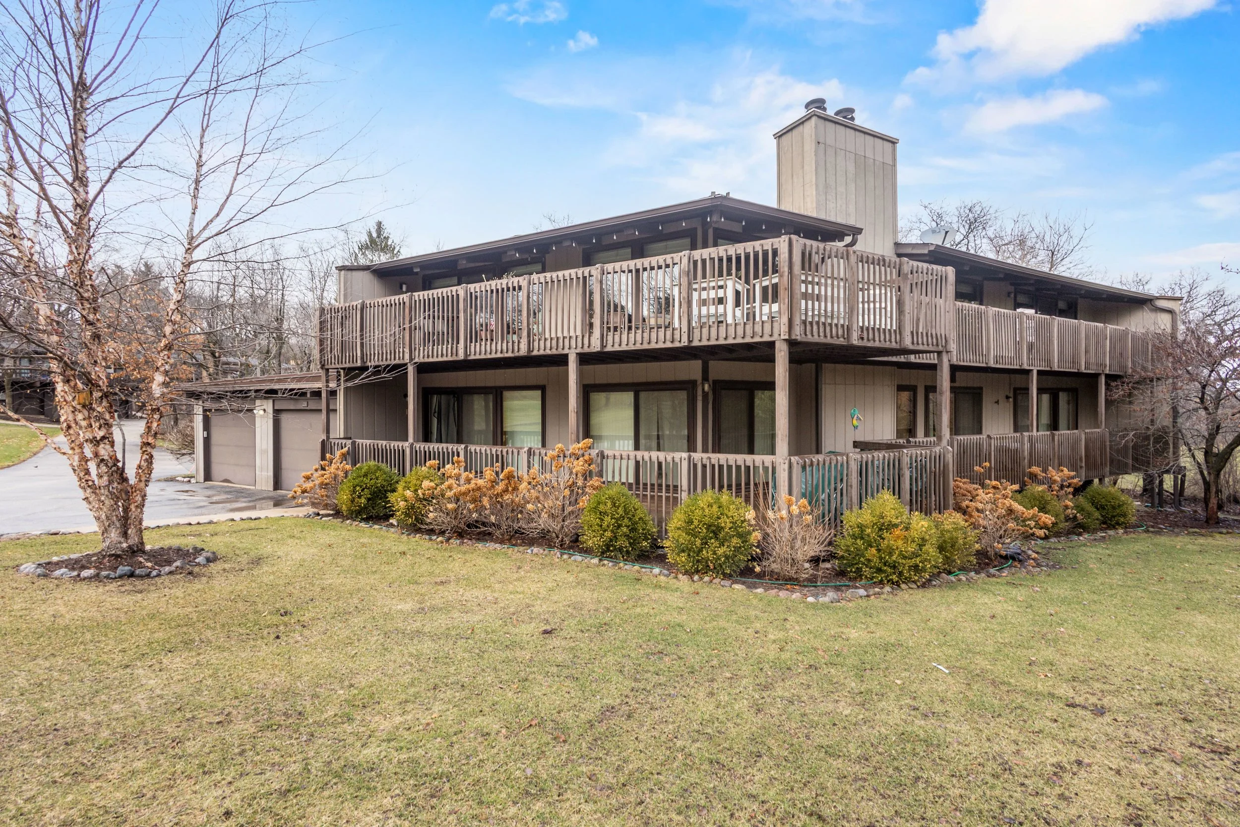 A two-story house with a large wooden deck and railing, surrounded by trees and bushes, under a partly cloudy sky.