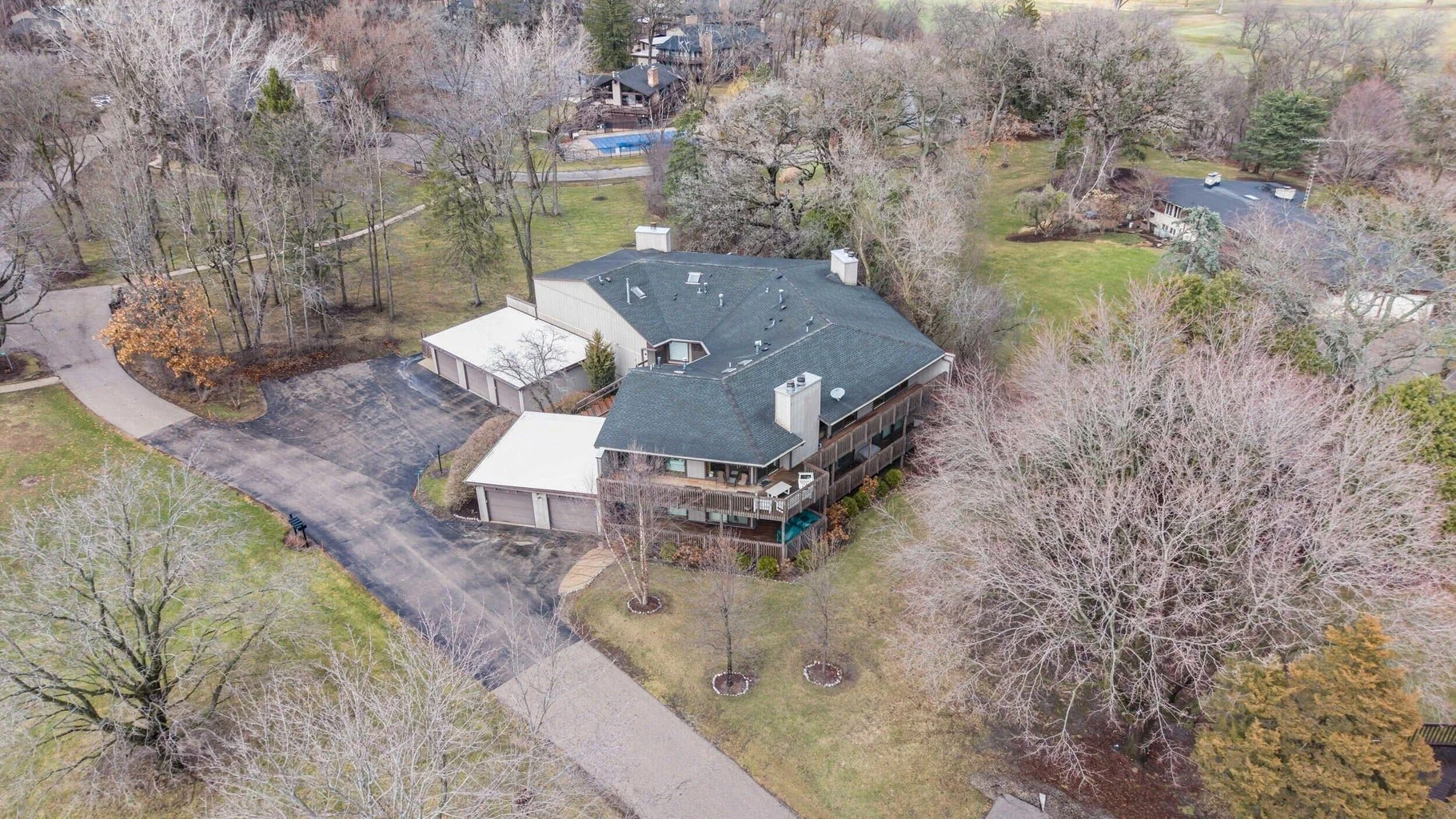 Aerial view of a residential house with a dark gray roof, surrounded by leafless trees, a driveway, and a backyard with a swimming pool and patio area.