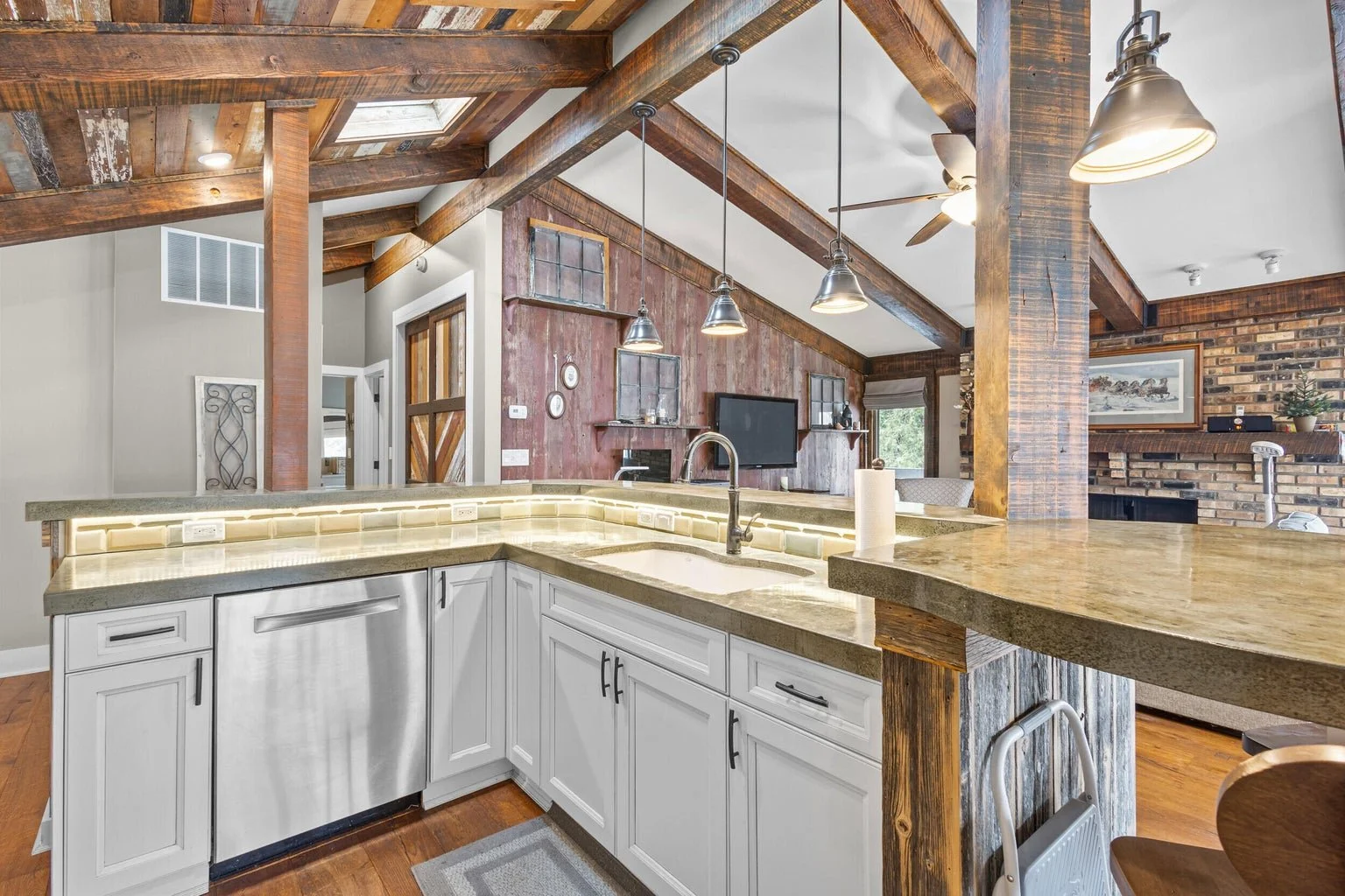 Open kitchen with white cabinets, stainless steel dishwasher, granite countertops, rustic wooden beams, pendant lights, and a view into the living room with a brick fireplace and wall-mounted television.