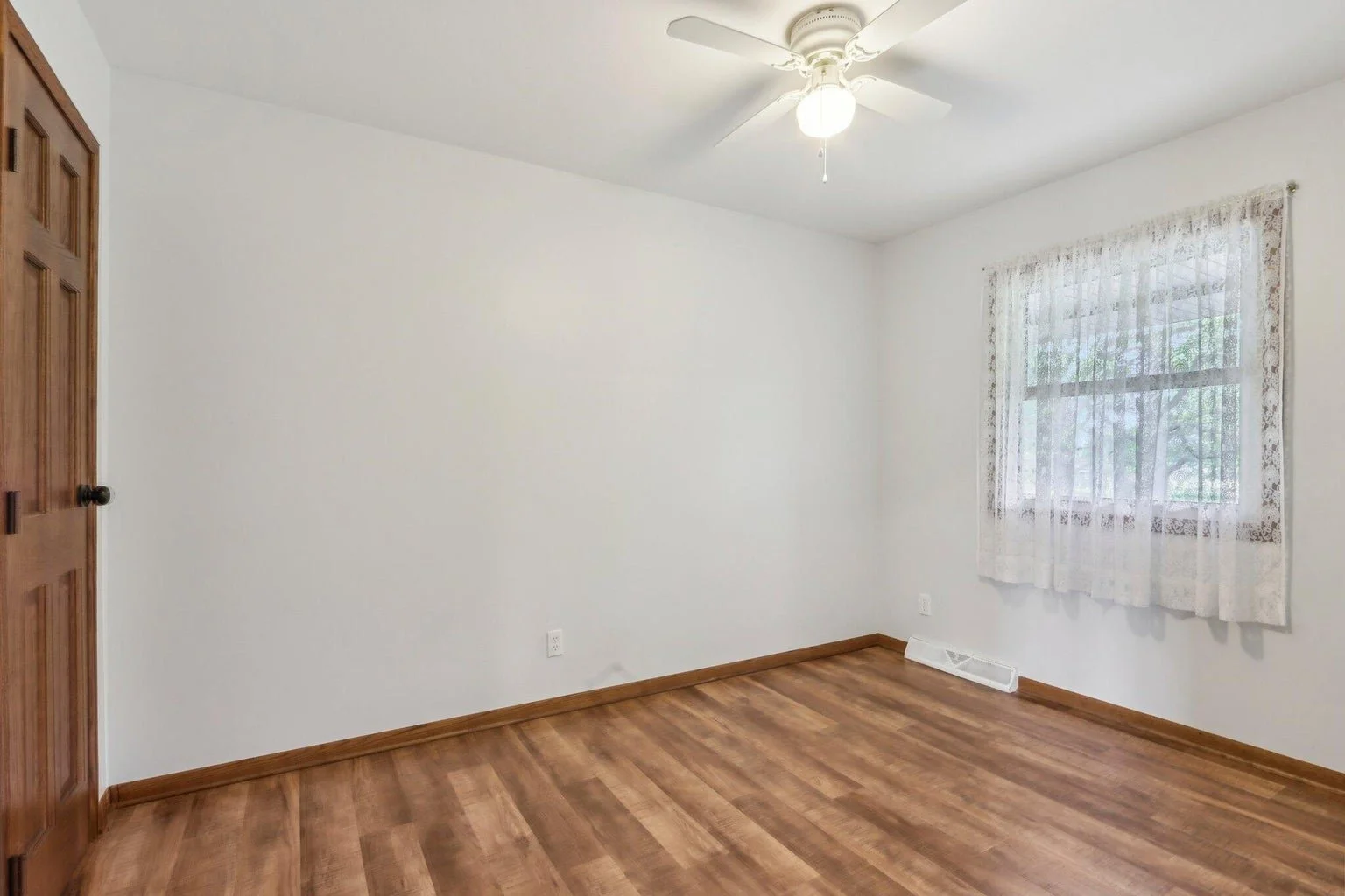 Empty room with wooden floors, white walls, a window with lace curtains, and a ceiling fan with a light.