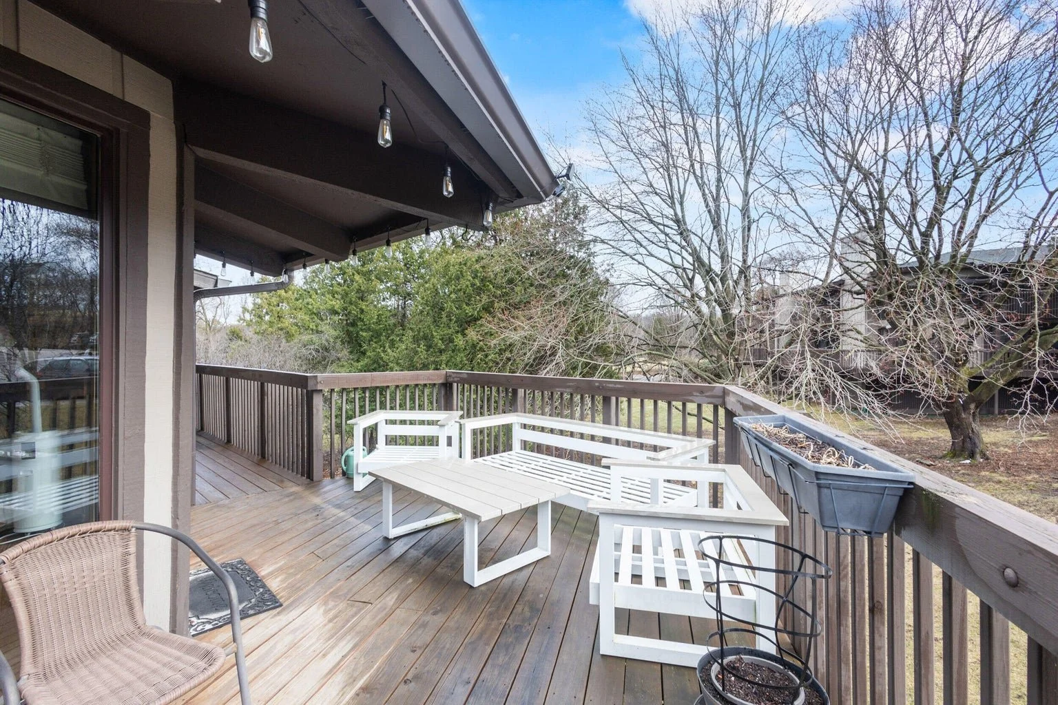 A wooden balcony with outdoor furniture, including white benches and a small table, overlooking a yard with trees.