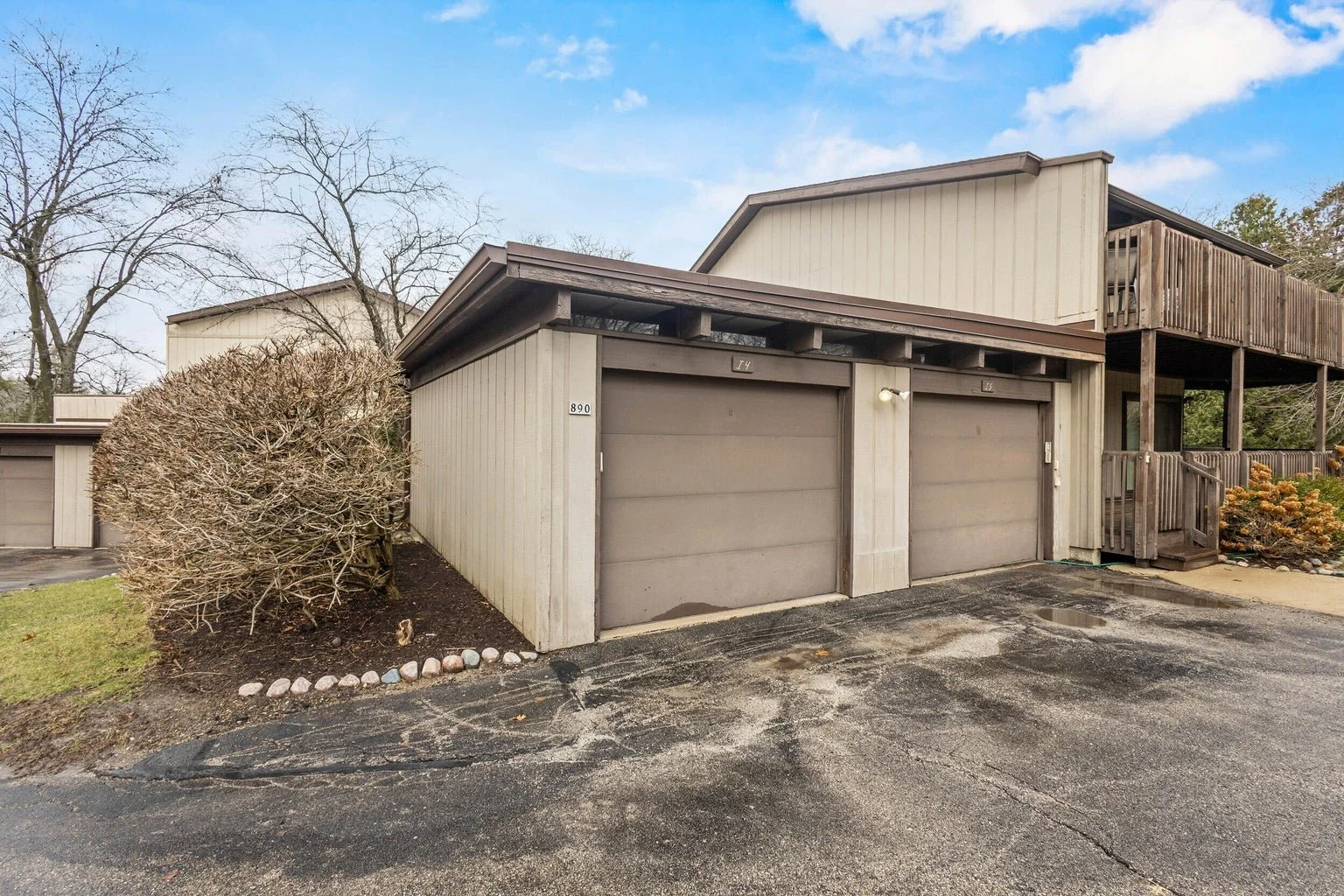 Exterior view of a beige residential building with attached two-car garage, a small porch with stairs, and a wooden balcony above. The driveway is cracked, and a leafless bush sits beside the garage. The sky is partly cloudy.