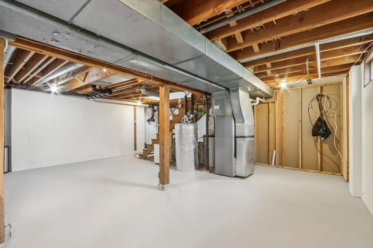 Empty basement with exposed wooden beams and HVAC ductwork, white painted walls, concrete floor, and some electrical wiring on the wall.