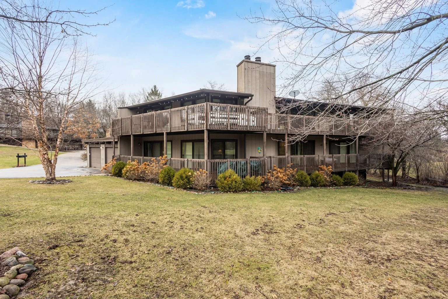 A two-story residential house with extensive wooden decks and a sloped backyard with grass, shrubs, and leafless trees, under a clear sky.