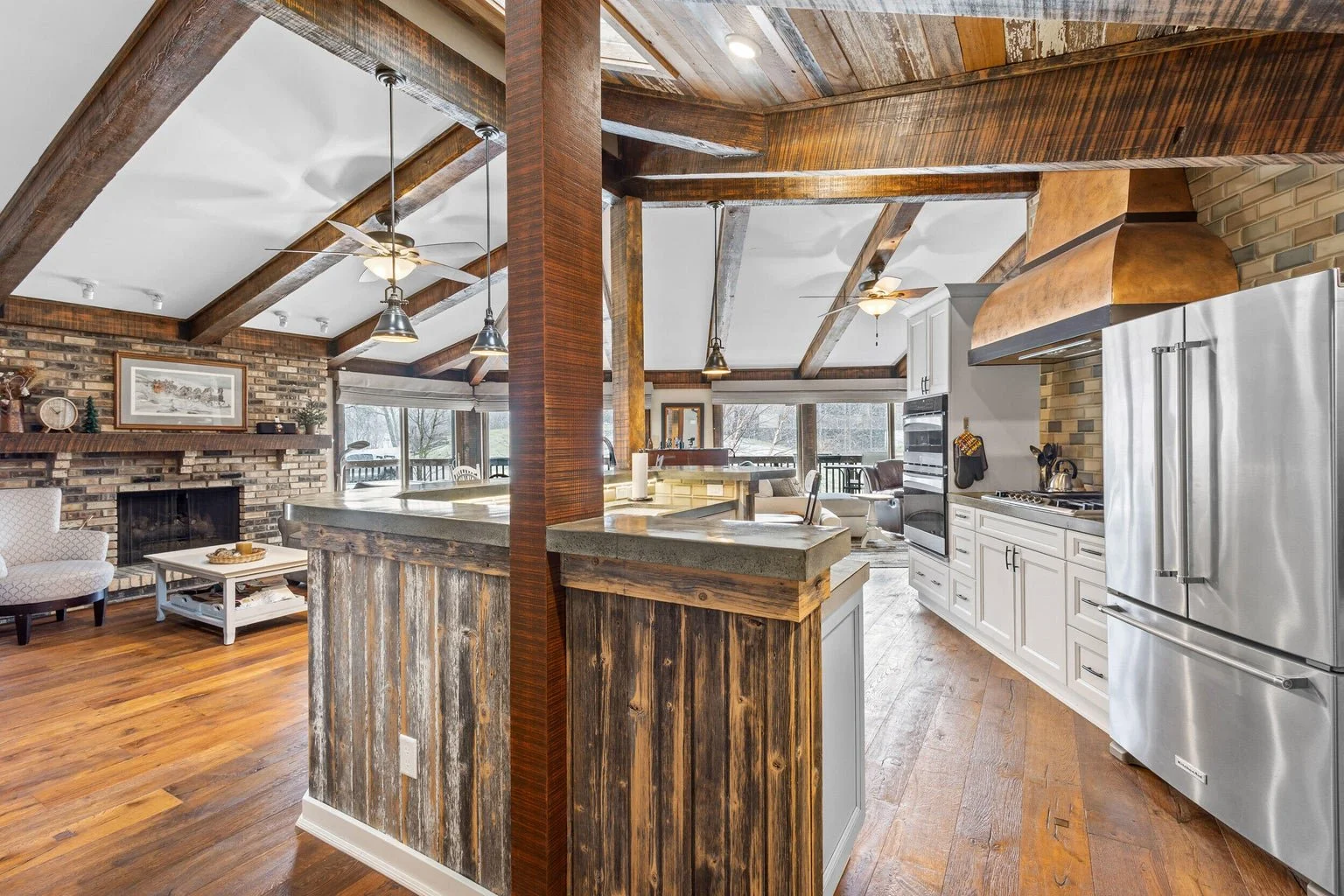 Open-concept kitchen and living area with exposed wooden beams, brick fireplace, white cabinets, stainless steel refrigerator, and a bar counter with rustic wood paneling.