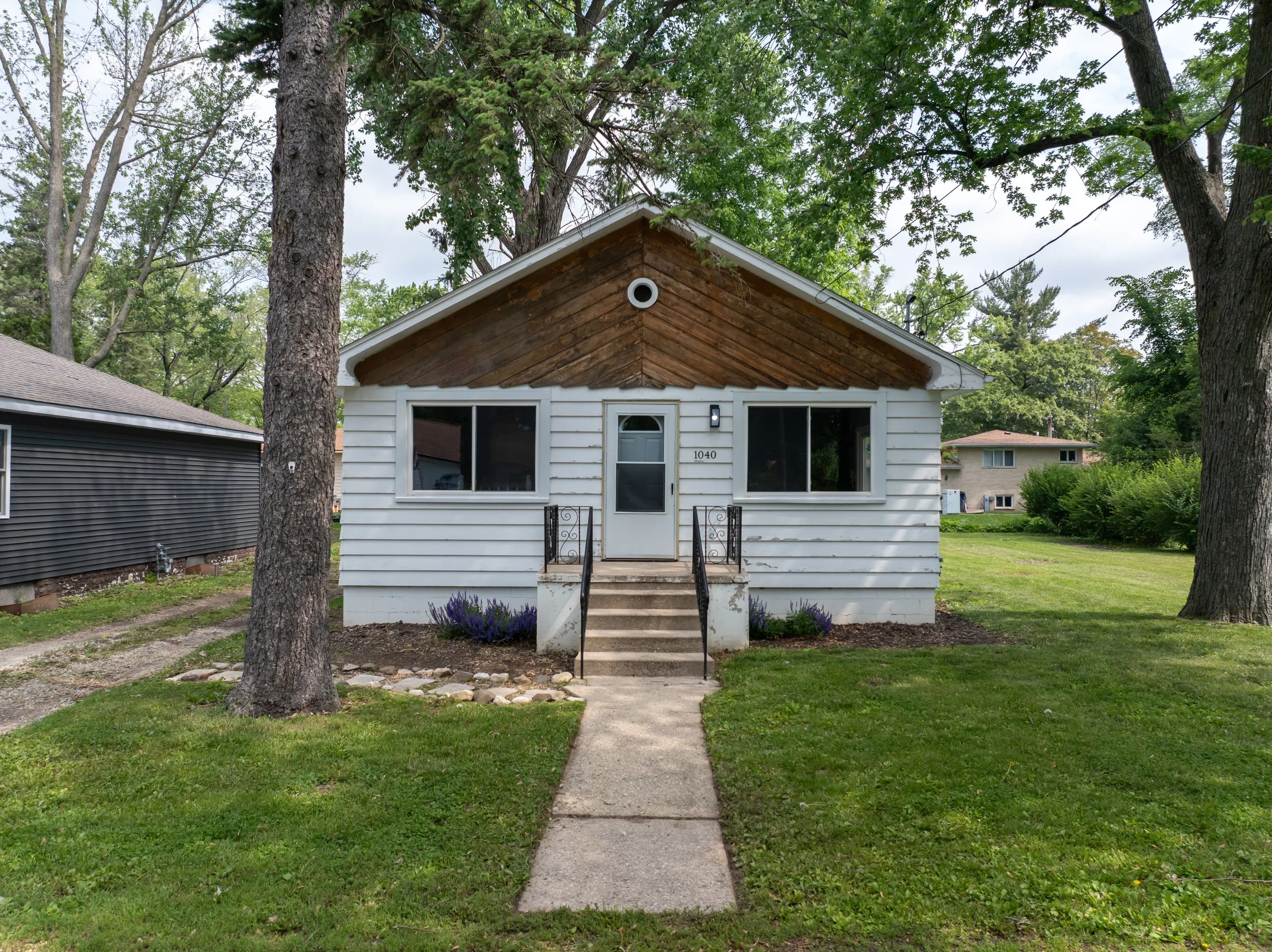 Front view of a small white house with a wooden front porch roof, three stairs, and a small front yard with a sidewalk. Large trees surround the house and neighboring houses are visible in the background.