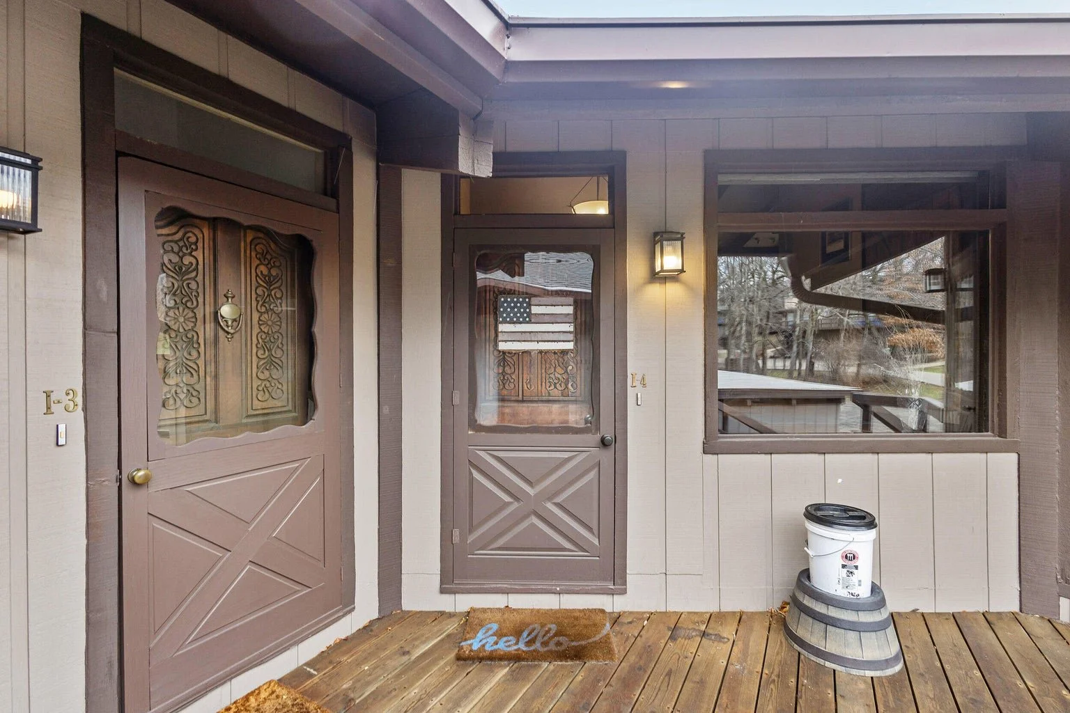 Front porch with two brown doors, one with glass and ornamental design, and a window to the right. A welcome mat says 'hello'. An American flag decoration is visible in the window. A bucket and a small barrel are on the wooden porch floor. Exterior w