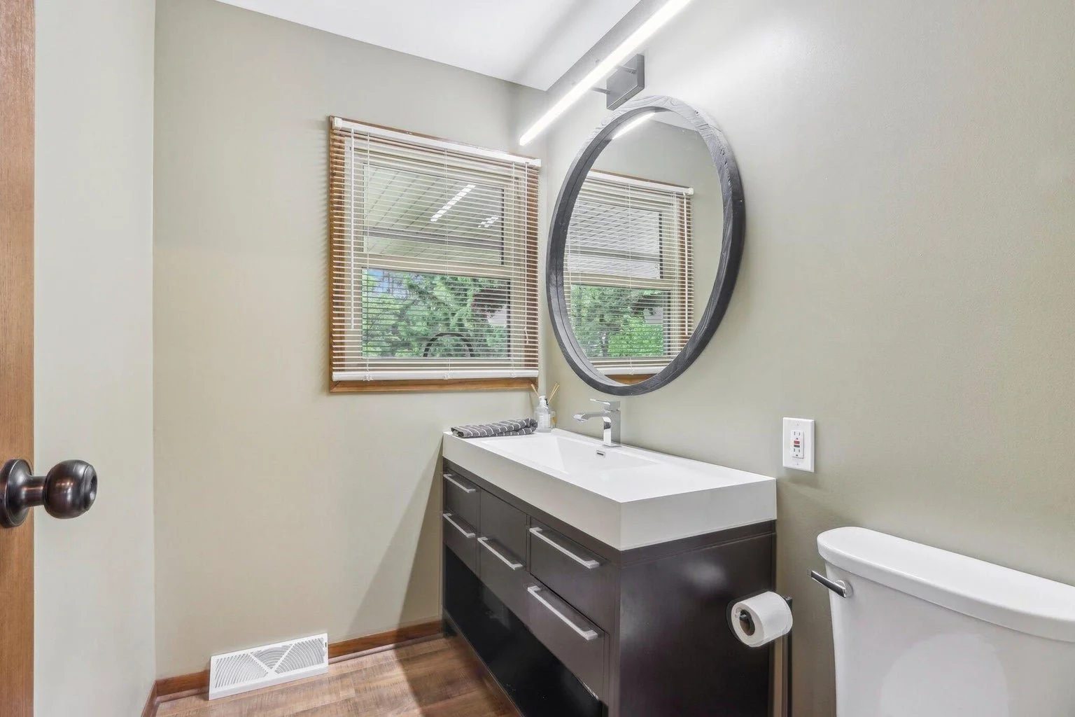 Bathroom with a white sink, dark vanity, large oval mirror, window with blinds, and a toilet with a toilet paper holder.