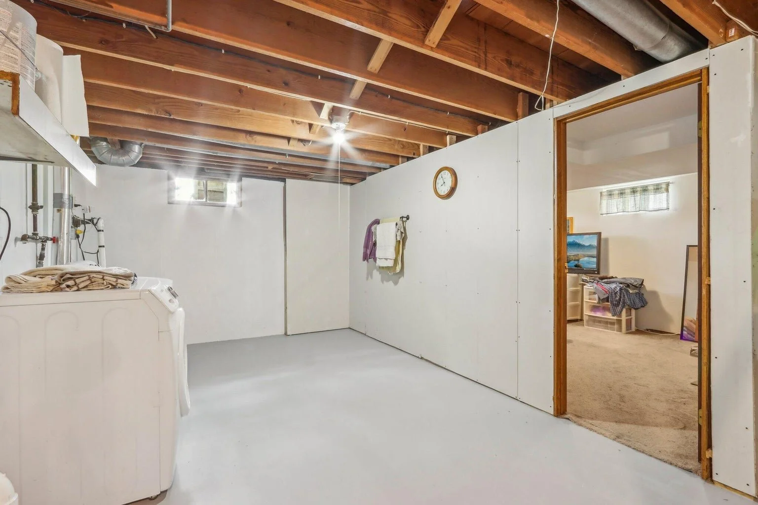 An unfinished basement with white painted concrete floors, exposed wooden ceiling beams, a small window, a wall clock, and a laundry area with a washing machine and towels hanging on a rack. An opening leads to a carpeted room with a television and a