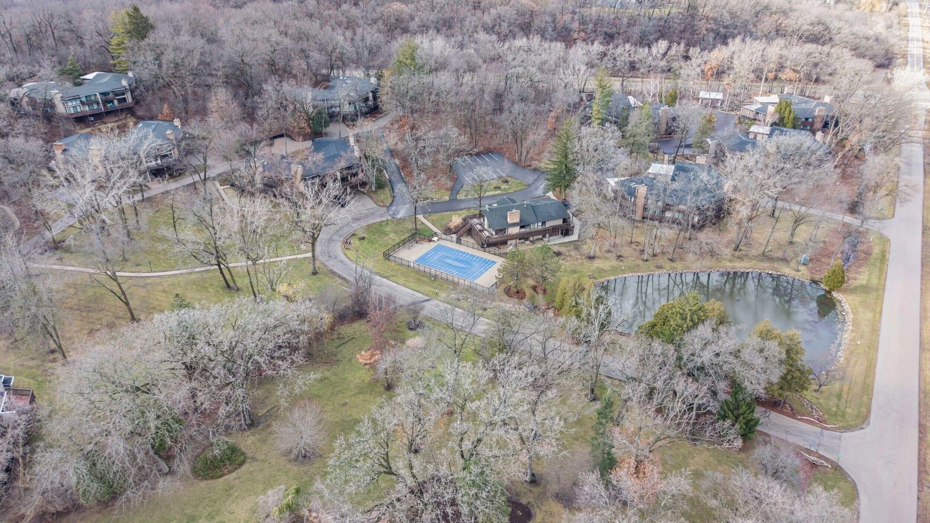 Aerial view of a residential area with multiple houses, trees, a small pond, and a tennis court surrounded by a fence. Roads and driveways connect the neighborhood.