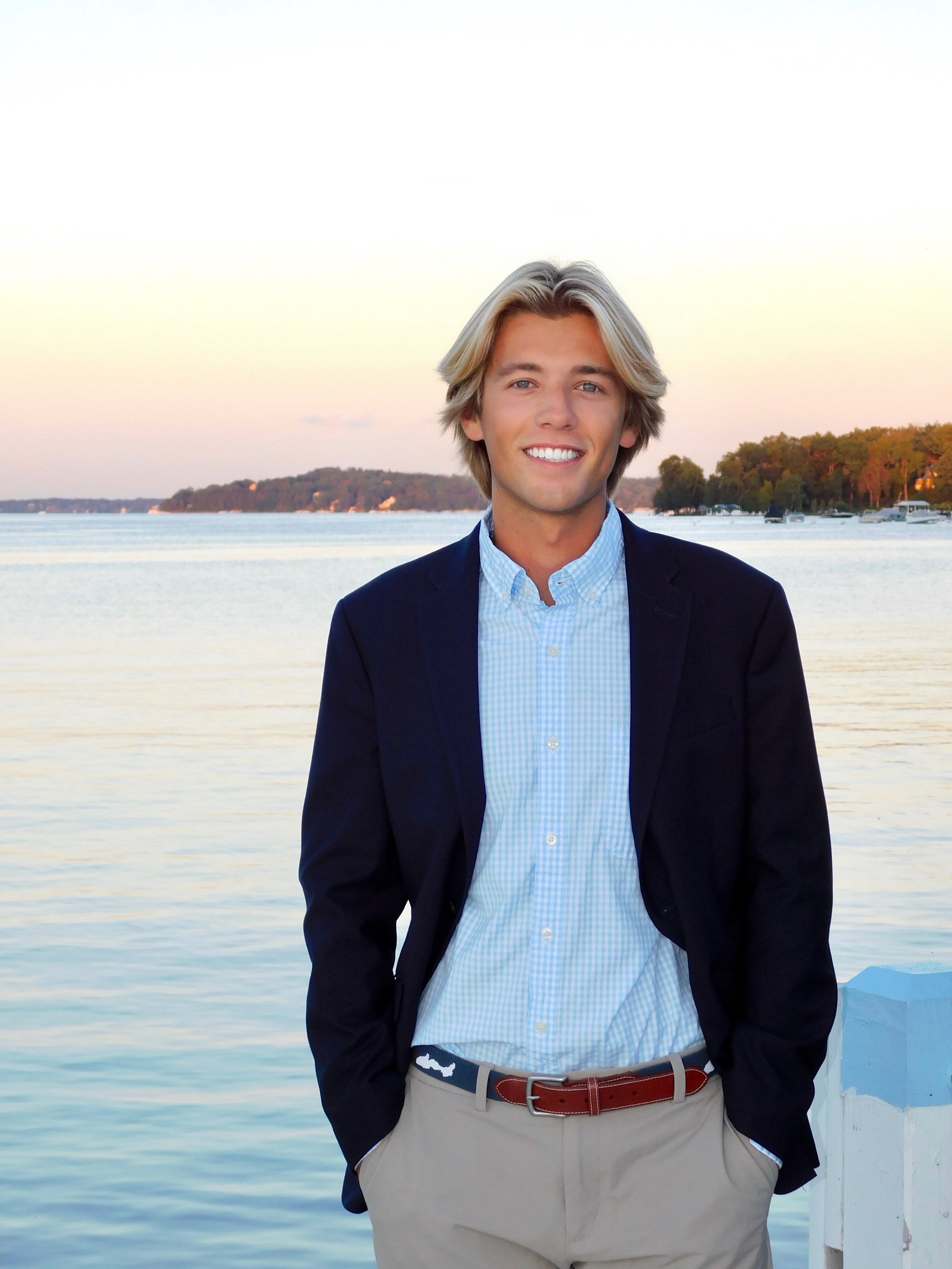 A young man in a navy blazer, light blue checkered shirt, and beige pants standing outdoors, smiling by a body of water during sunset.