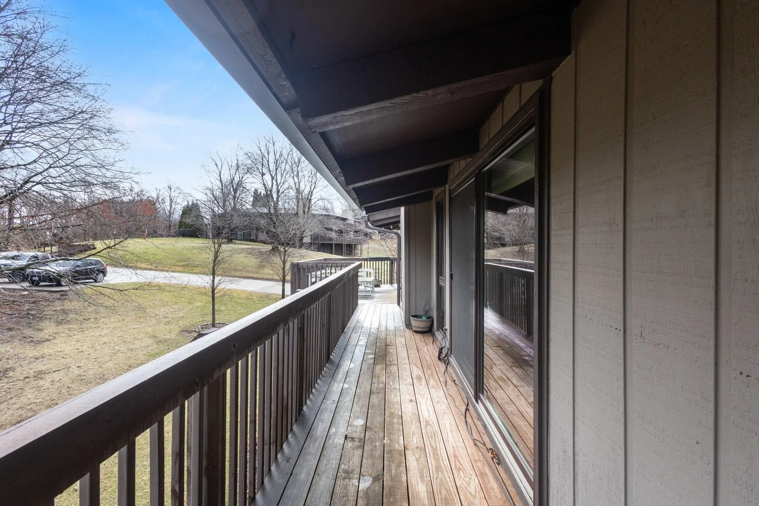 View from a wooden balcony with a sliding glass door, overlooking a grassy yard with leafless trees and parked cars, under a cloudy sky.