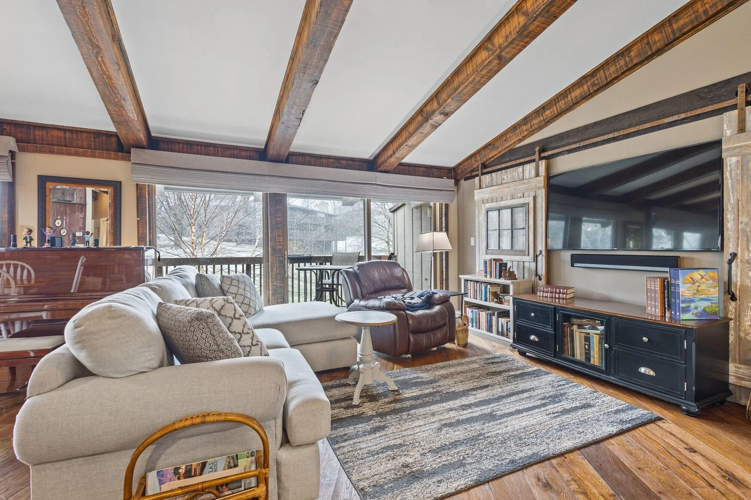 Living room with beige sectional sofa, leather armchair, bookshelf, large TV, large window with snow outside, and wooden ceiling beams