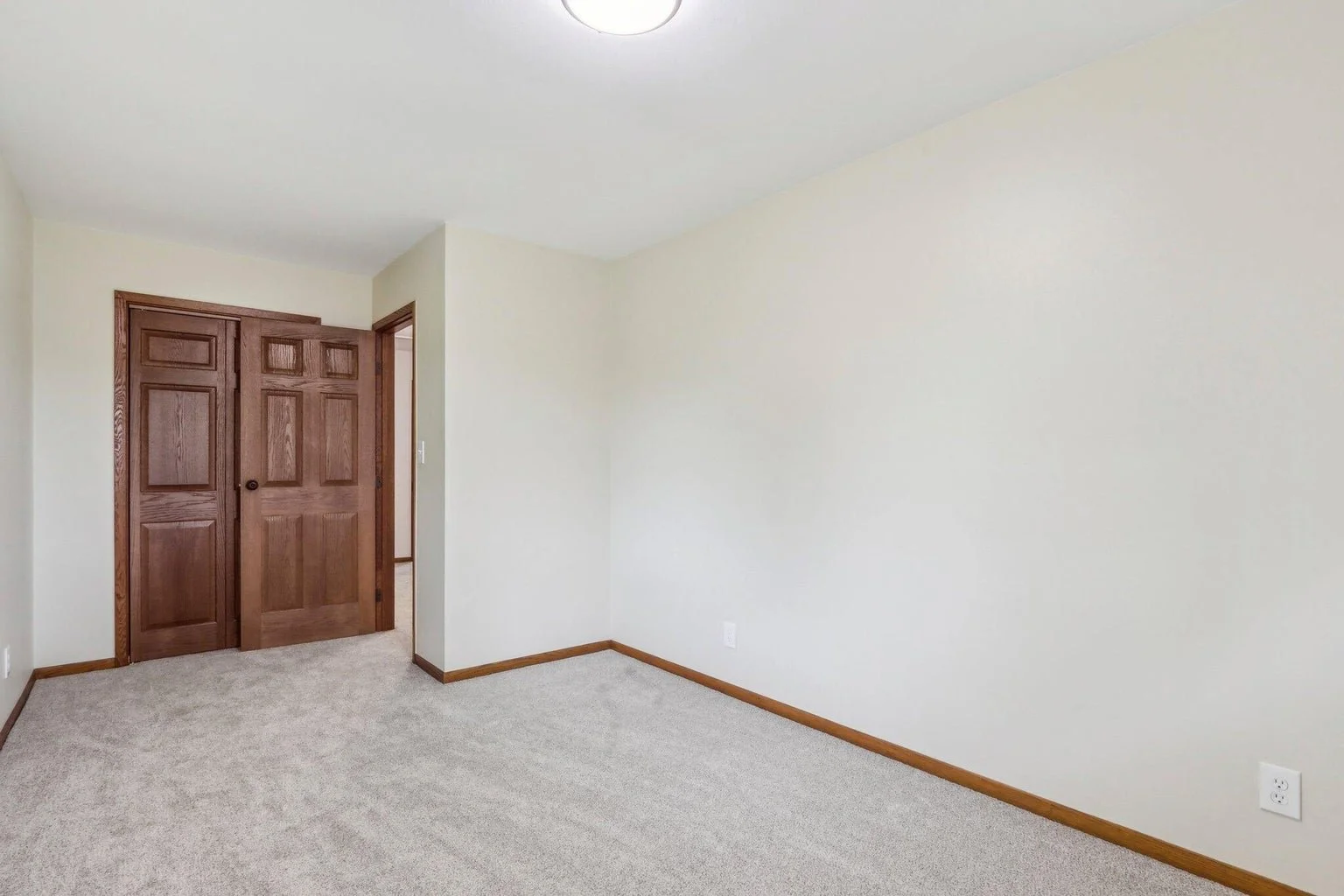 Empty room with beige carpet, cream-colored walls, and wooden trim. There are double closet doors made of dark wood, and a white ceiling with a round light fixture.