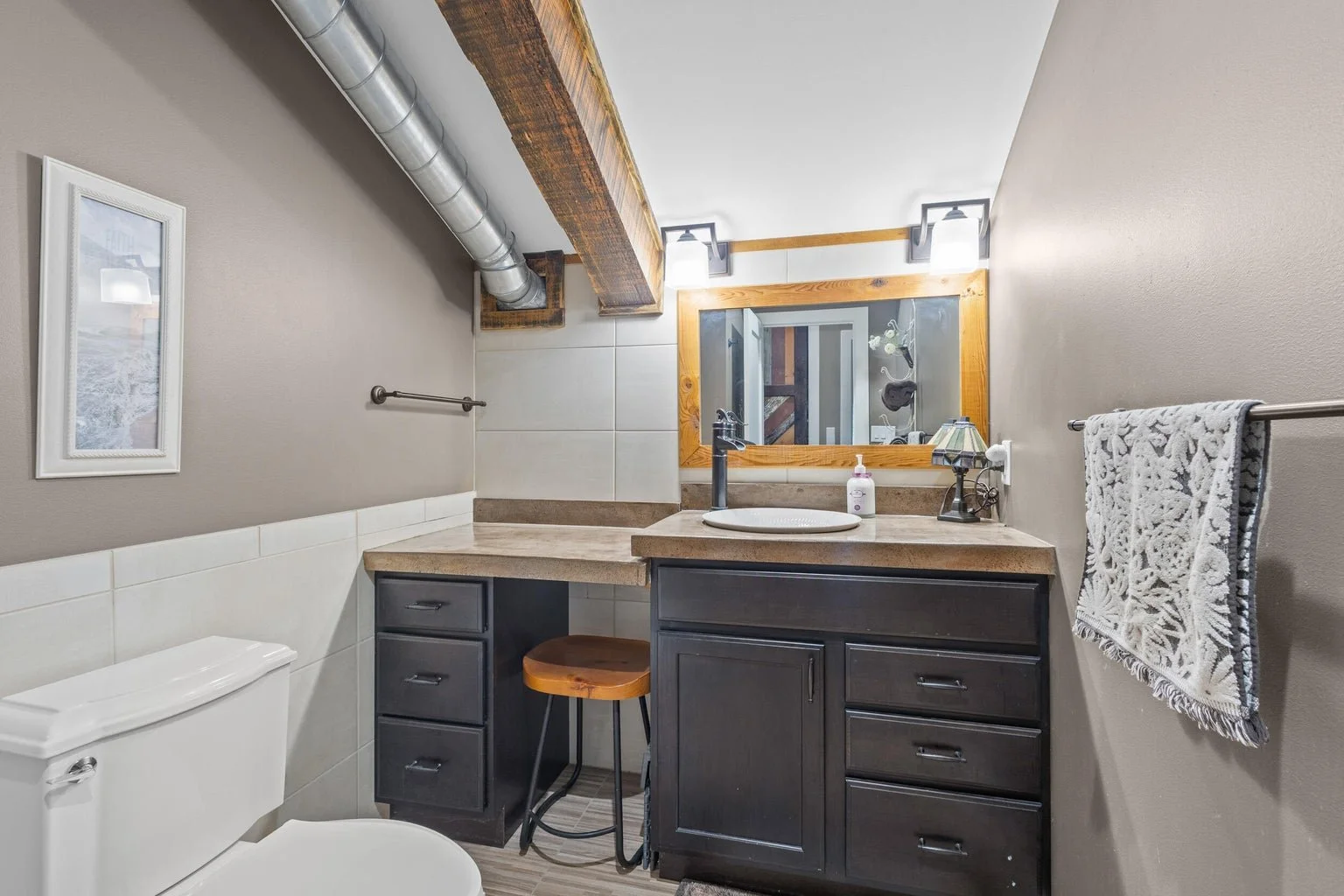 Small bathroom with a neutral color palette, featuring a white toilet, dark wooden vanity with a beige countertop, a round sink, a large mirror with a wooden frame, and a patterned towel hanging on a metal rod. Exposed ductwork and wooden beams are v