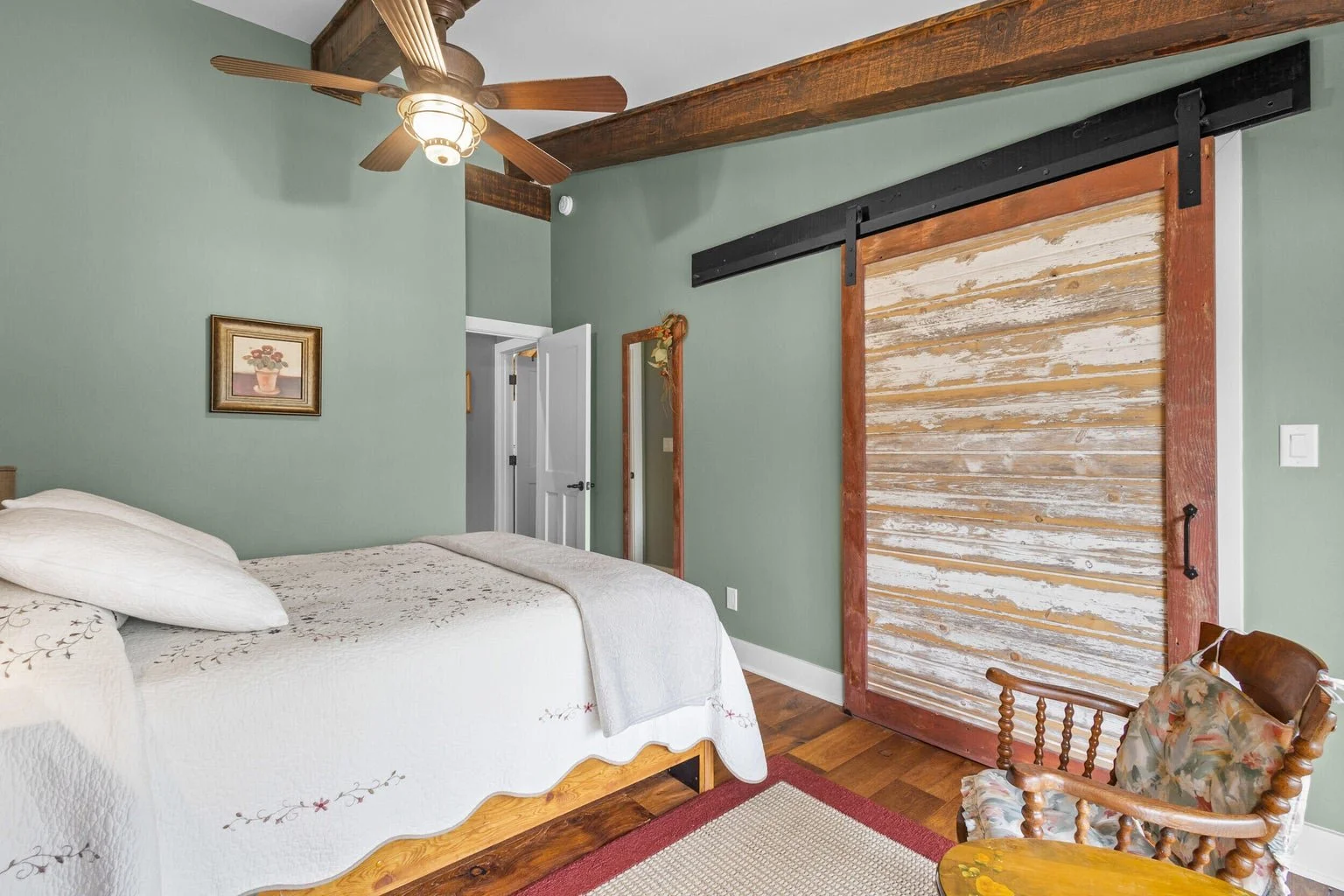 A bedroom with mint green walls, a ceiling fan, a bed with white quilted bedding, a wooden chair with floral cushion, a small table, and a rustic sliding barn door made of weathered wood with red trim.