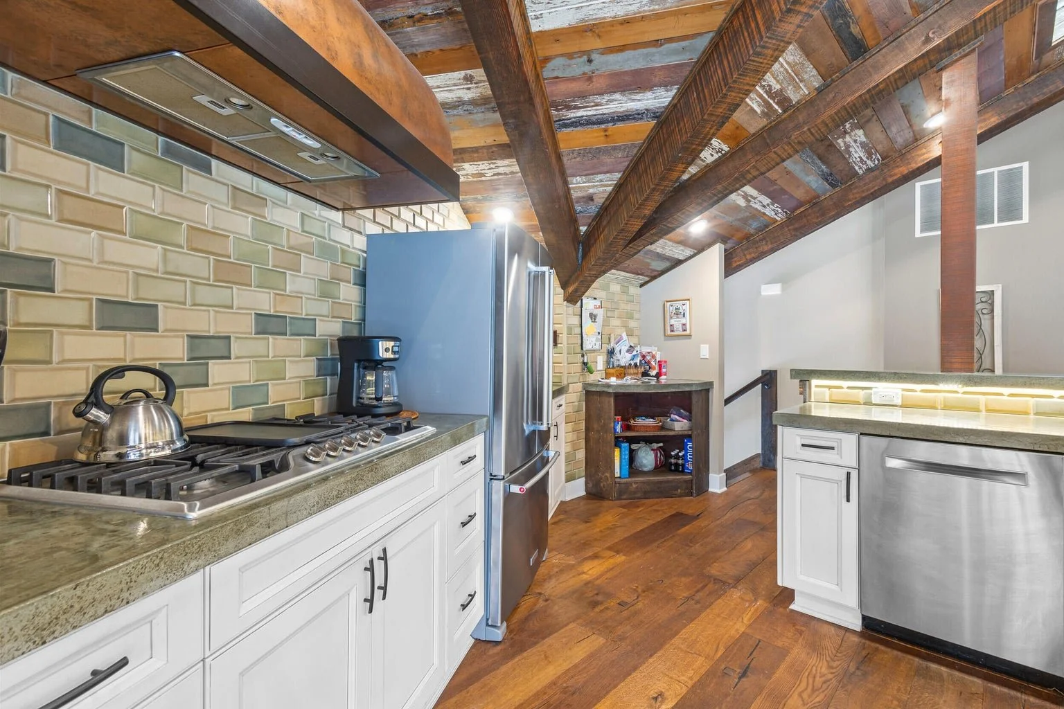 A kitchen with wooden ceiling beams, a brick backsplash, white cabinets, stainless steel appliances, and hardwood flooring.