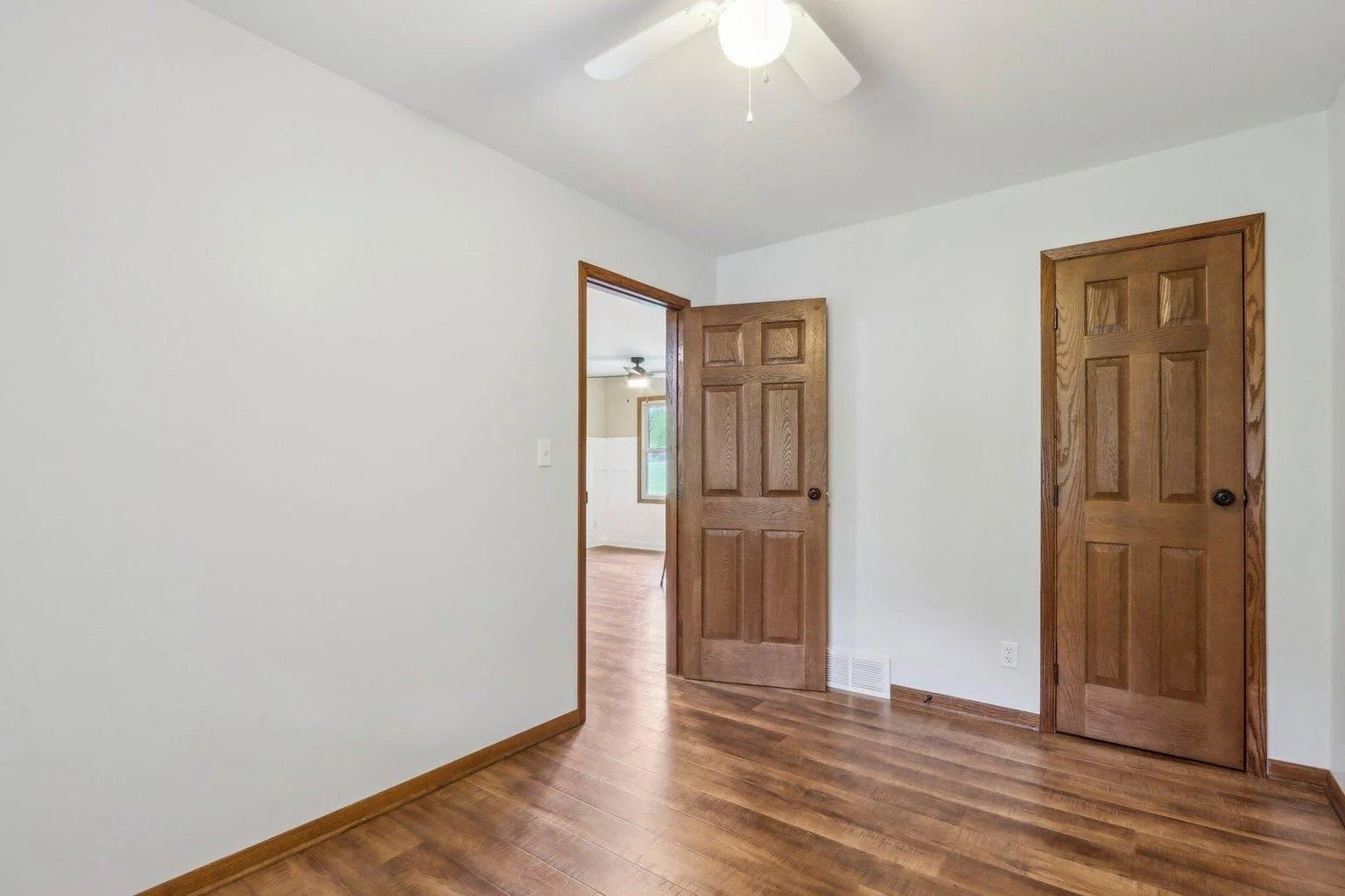 Empty room with hardwood floors, white walls, wooden trim, ceiling fan with light, and two closed wooden doors.