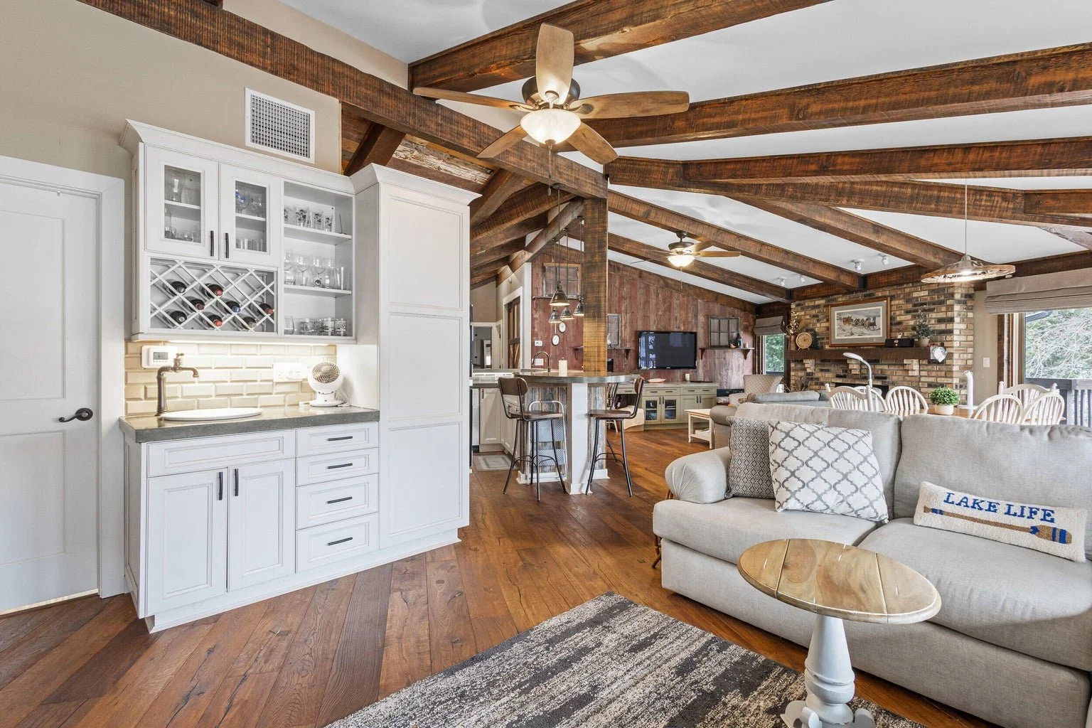 Living room with wood beamed ceiling, white sofa with decorative pillows, small wooden side table, and a kitchen area with a white cabinet, bar with stools, and a brick fireplace with a painting above it, large windows, and hardwood floors.