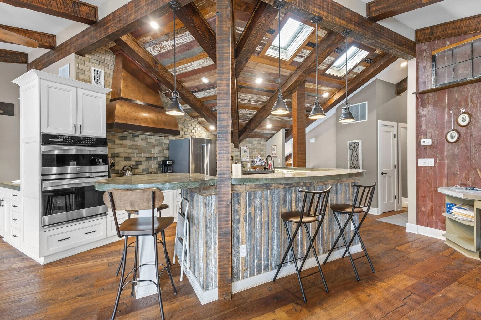 Kitchen with rustic wood and metal accents, white cabinets, stainless steel appliances, a large copper hood, a curved island with a wood-paneled exterior, black barstools, pendant lighting, hardwood floors, and vaulted ceilings with exposed wood beam