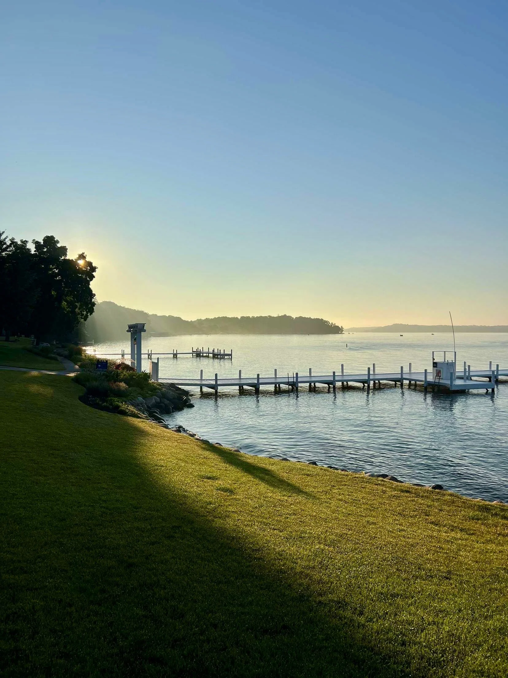 A peaceful lakeside scene during sunrise or sunset with a grassy shore, trees on the left, a dock extending into the water, and distant hills on the horizon.