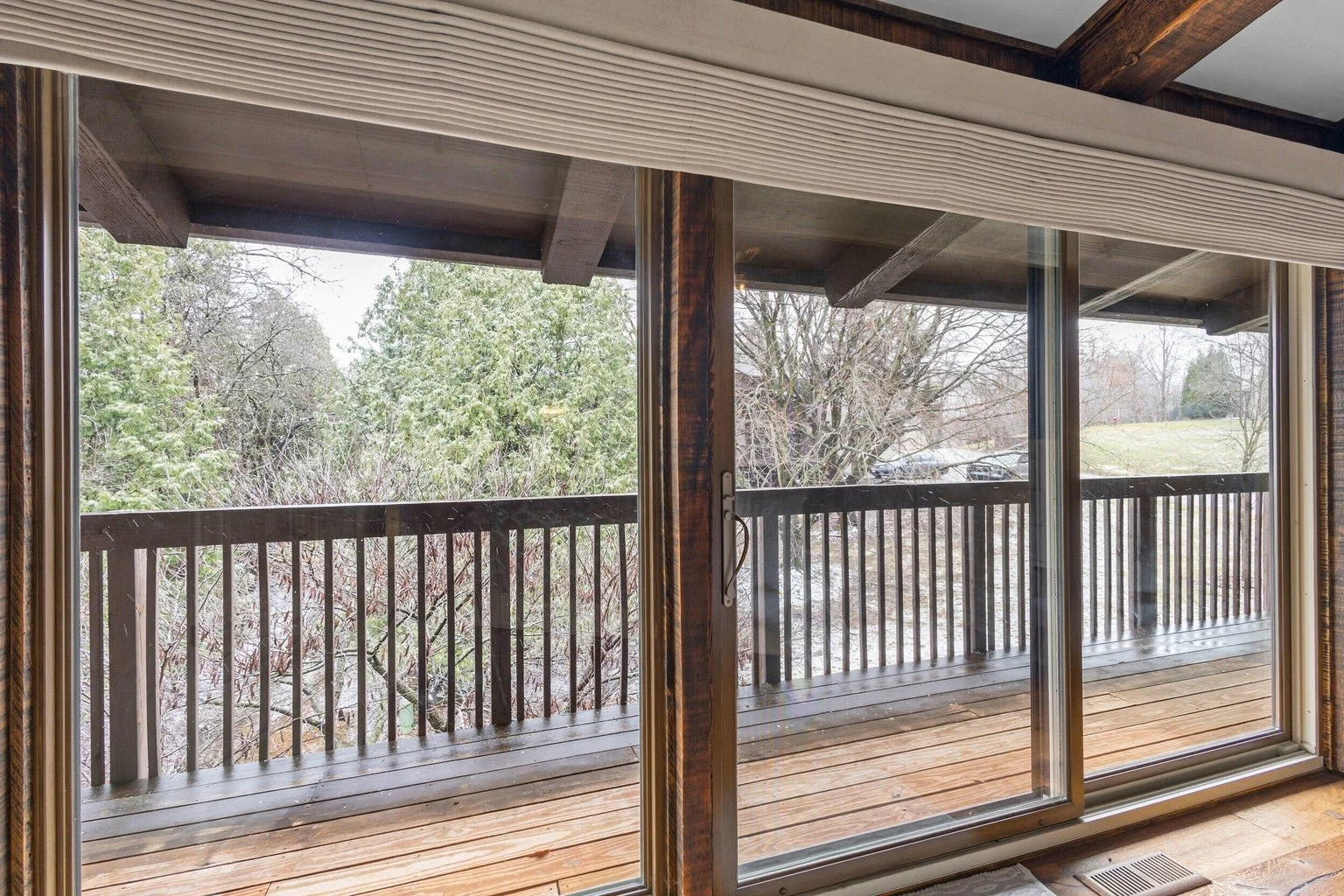 View from inside a house looking out through a glass sliding door at a wooden balcony with a railing, overlooking trees and a grassy area.