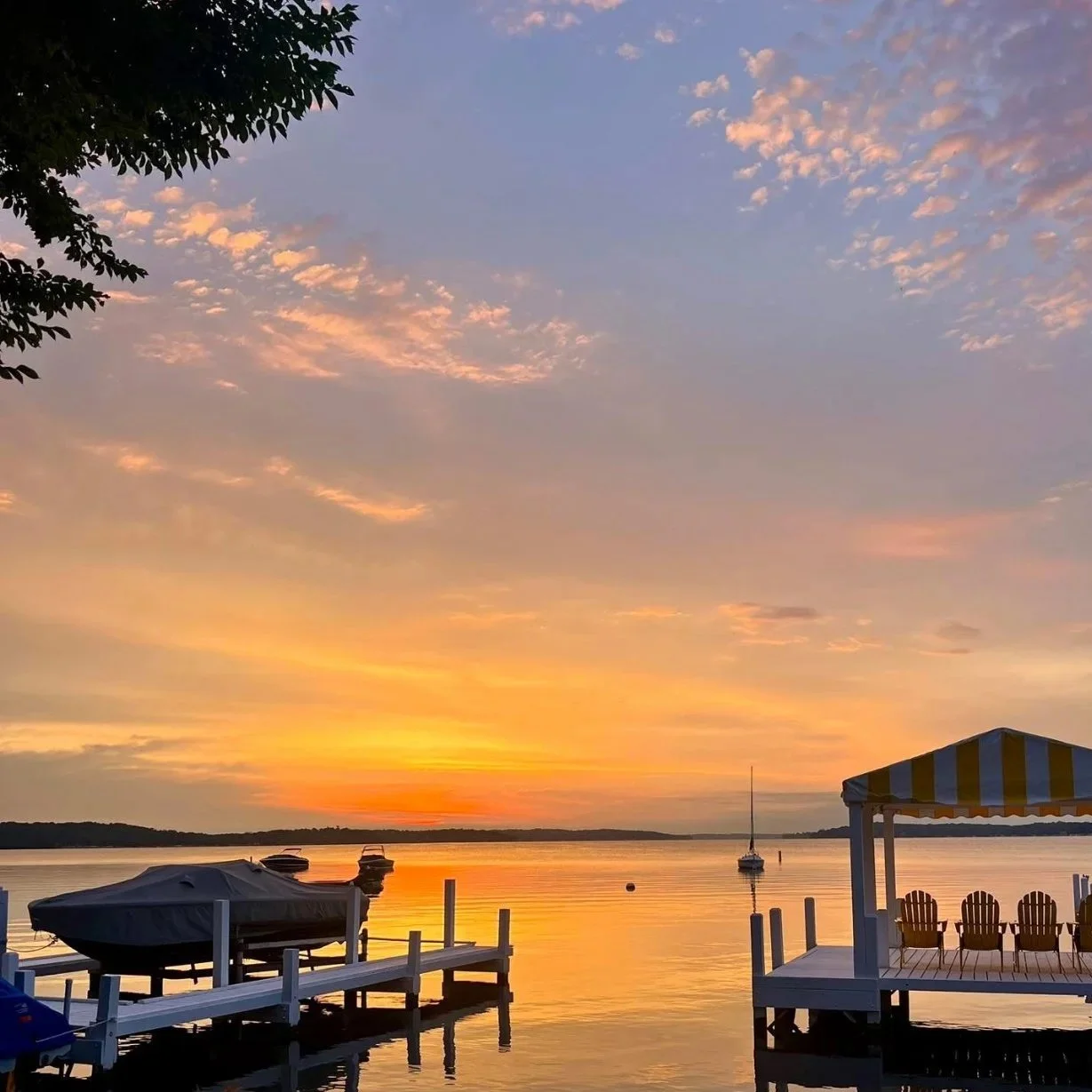A sunset over a calm body of water with boats docked by a pier and a gazebo on the right, featuring a colorful striped roof and outdoor chairs, with the sky filled with pink and orange clouds.
