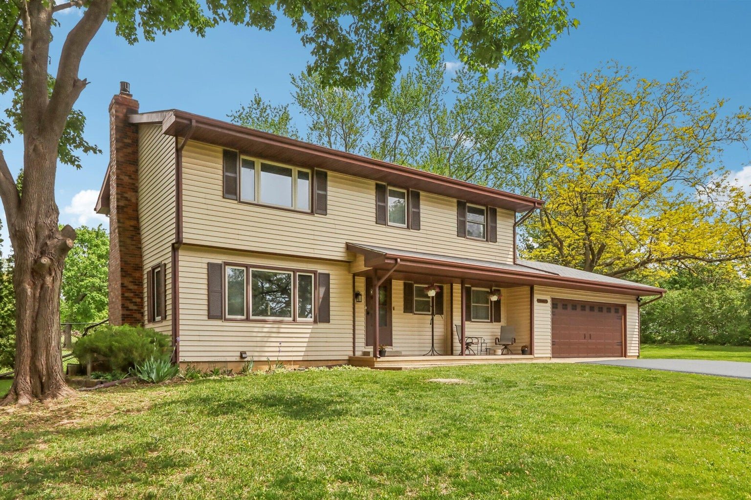 A two-story suburban house with beige siding, brown shutters, and a brown garage door, surrounded by green trees and a well-maintained lawn.