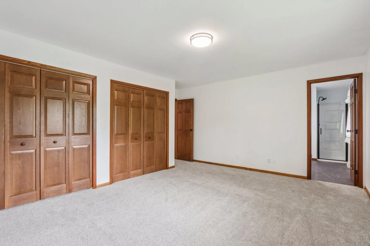 Empty bedroom with beige carpet, white walls, and wooden doors including closet doors and a doorway leading to a bathroom with a shower.