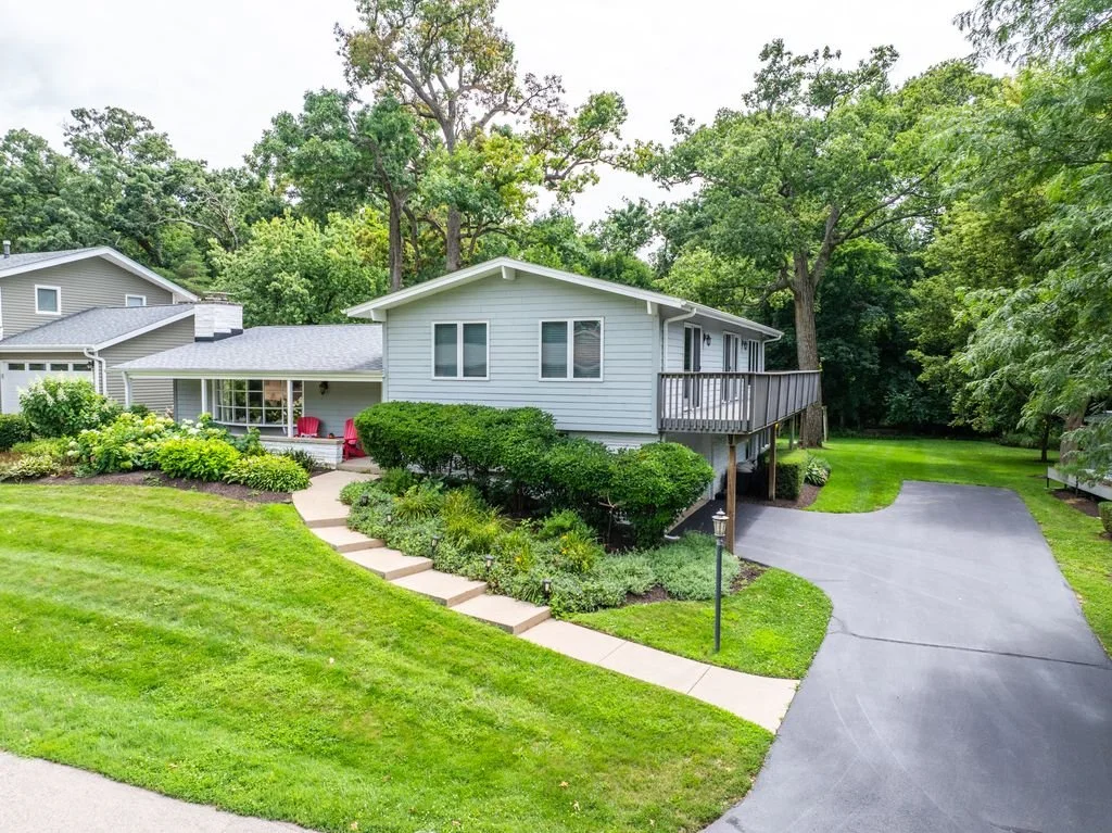 A suburban house with a raised deck, surrounded by lush green trees and a well-maintained lawn with a curved driveway.