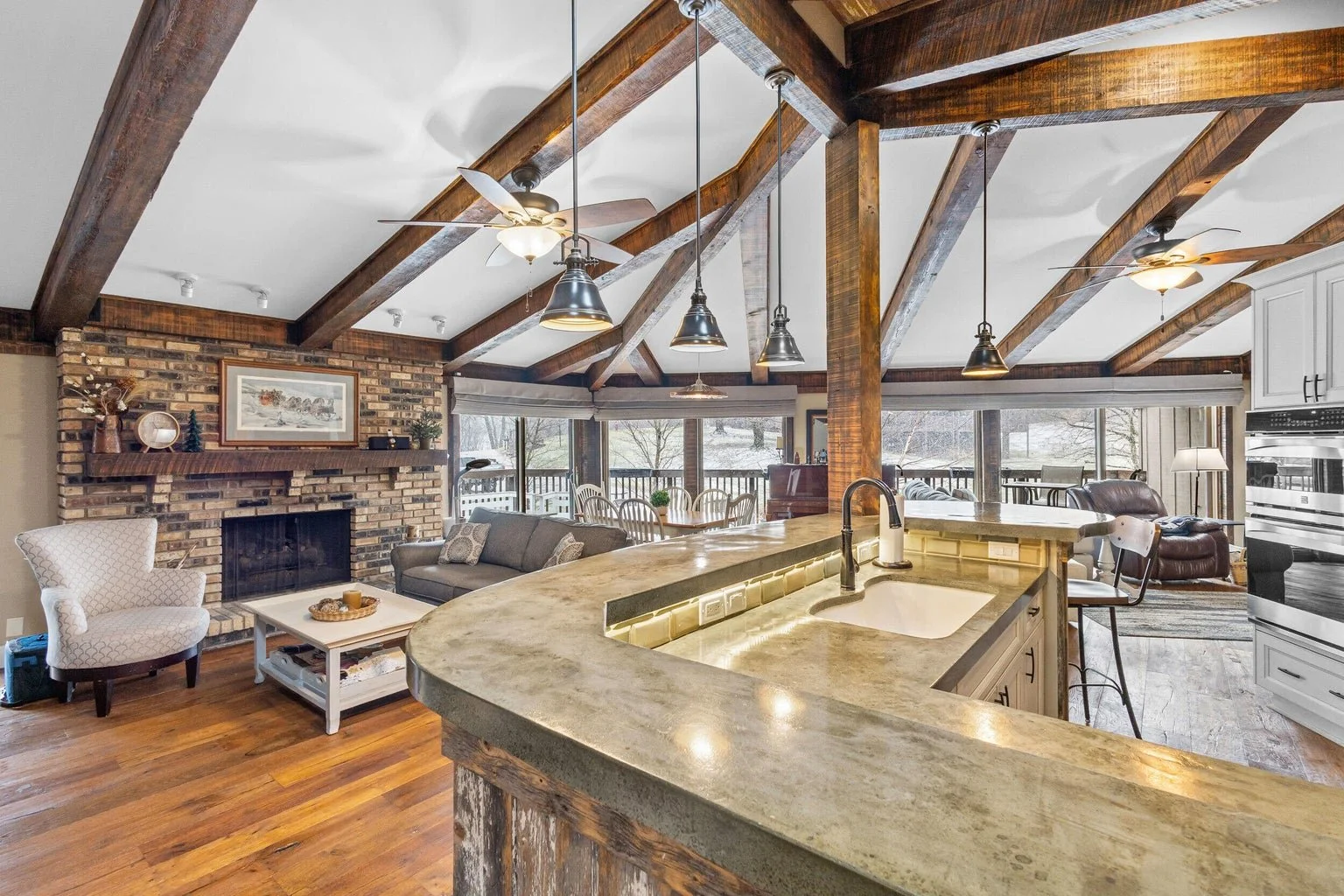 Open-concept living room and kitchen with wooden beams, brick fireplace, large windows, and casual furniture.