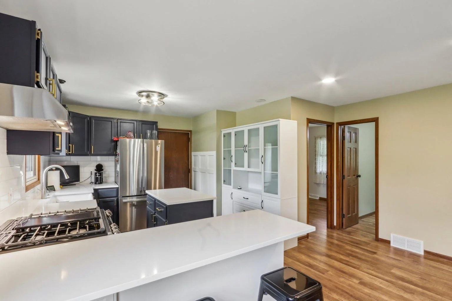 Modern kitchen with black cabinets, stainless steel refrigerator, white countertops, wood floors, and a white glass-front cabinet.