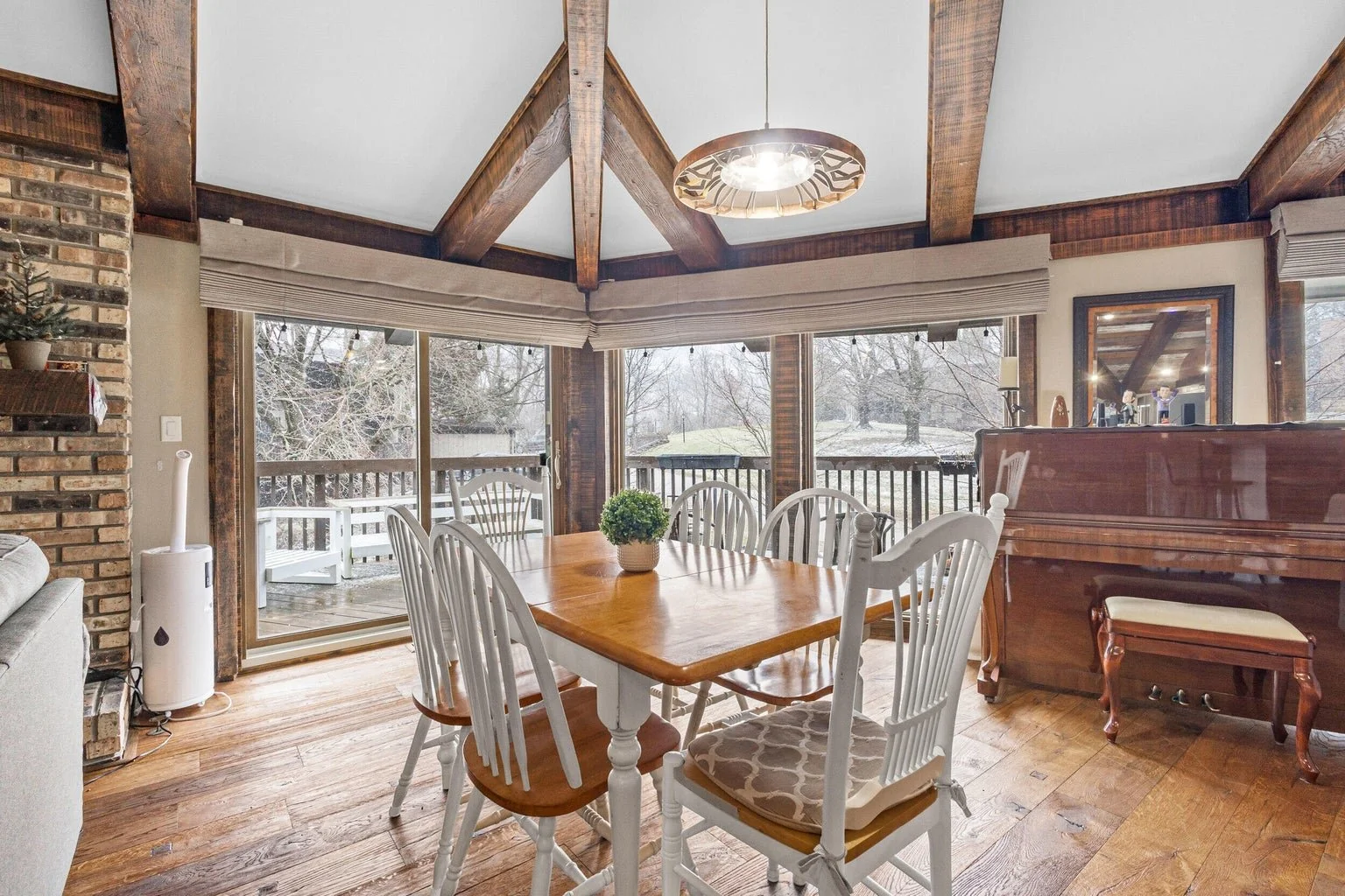 Dining room with a wooden table surrounded by white chairs, a piano, large window with a view of a snowy yard, and wooden beams on the ceiling.