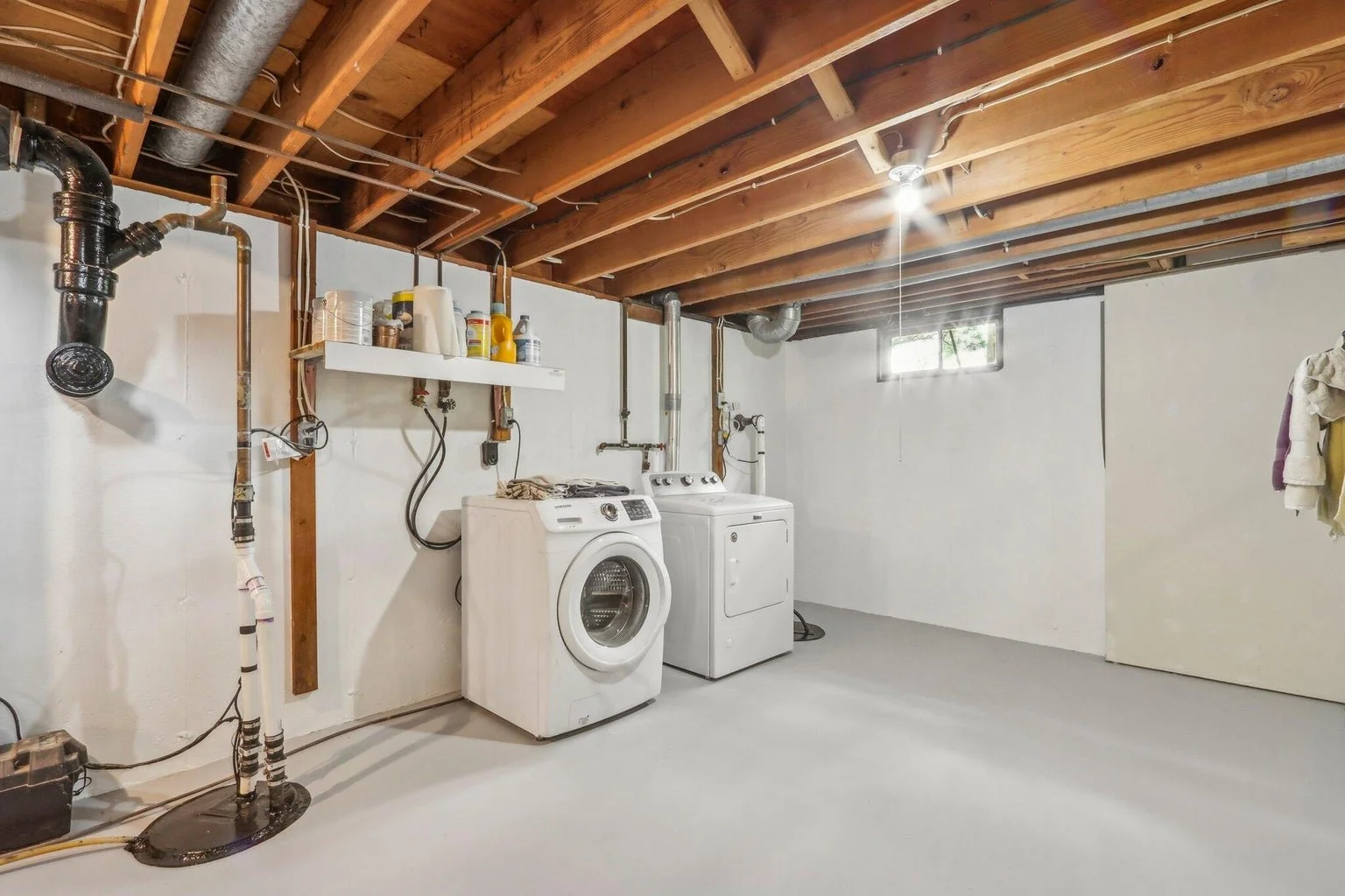Laundry room with a washing machine, dryer, laundry supplies on a white shelf, white walls, and an unfinished ceiling with exposed wooden beams and pipes.