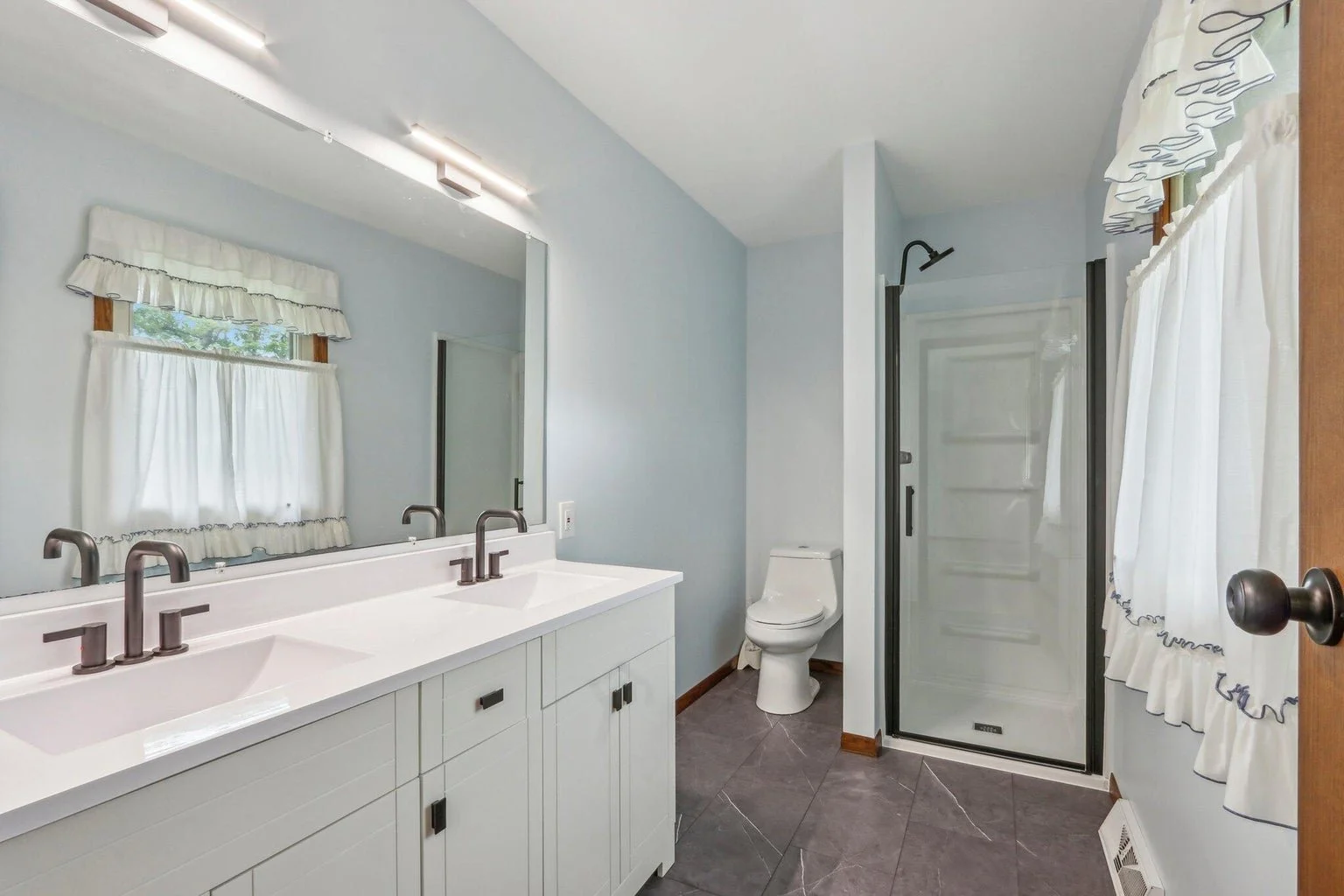 Bathroom featuring a double sink vanity, large mirror, window with white curtains, toilet, shower with glass door, and light blue walls.