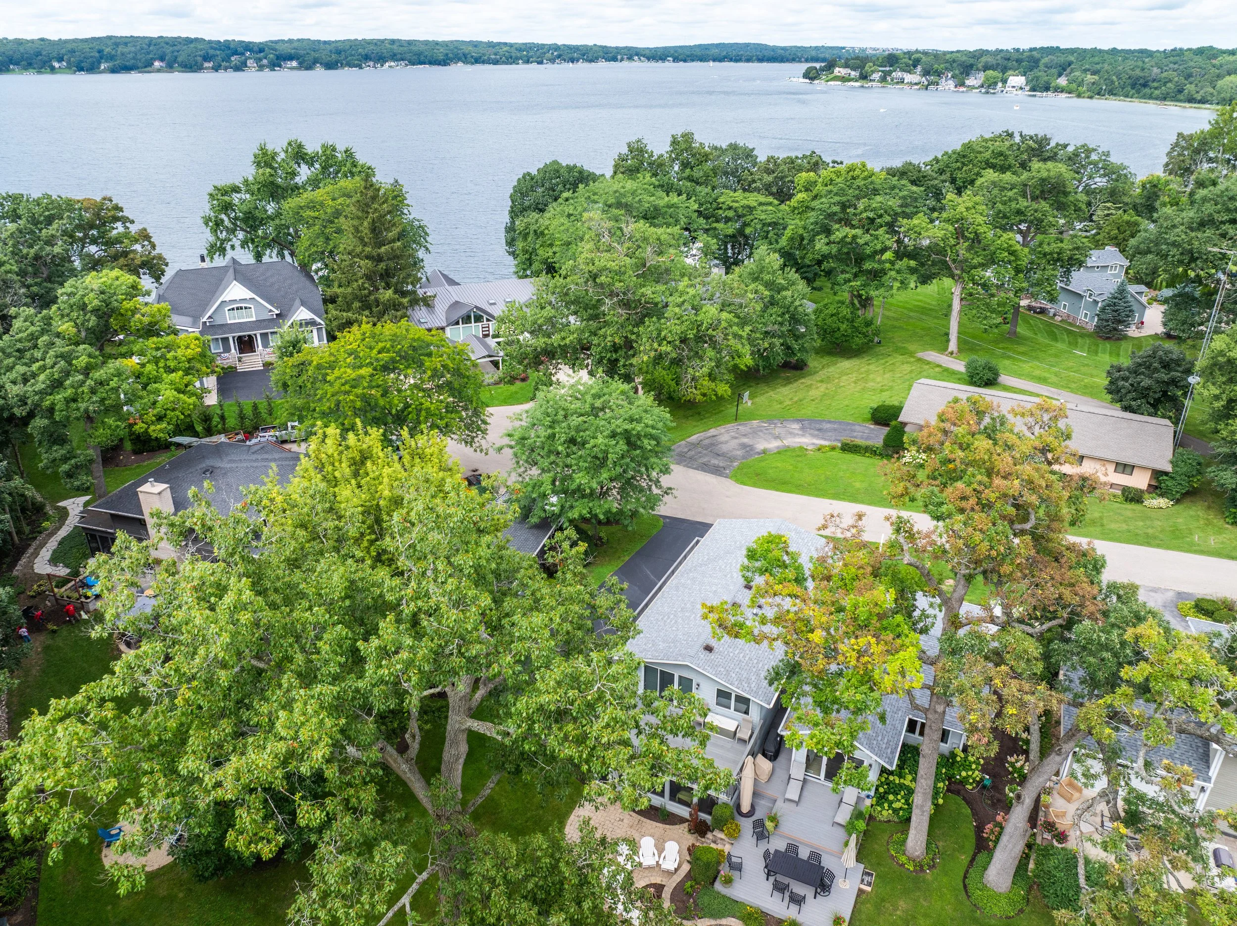 Aerial view of lakeside houses surrounded by trees and green lawns, with a large lake in the background under a partly cloudy sky.
