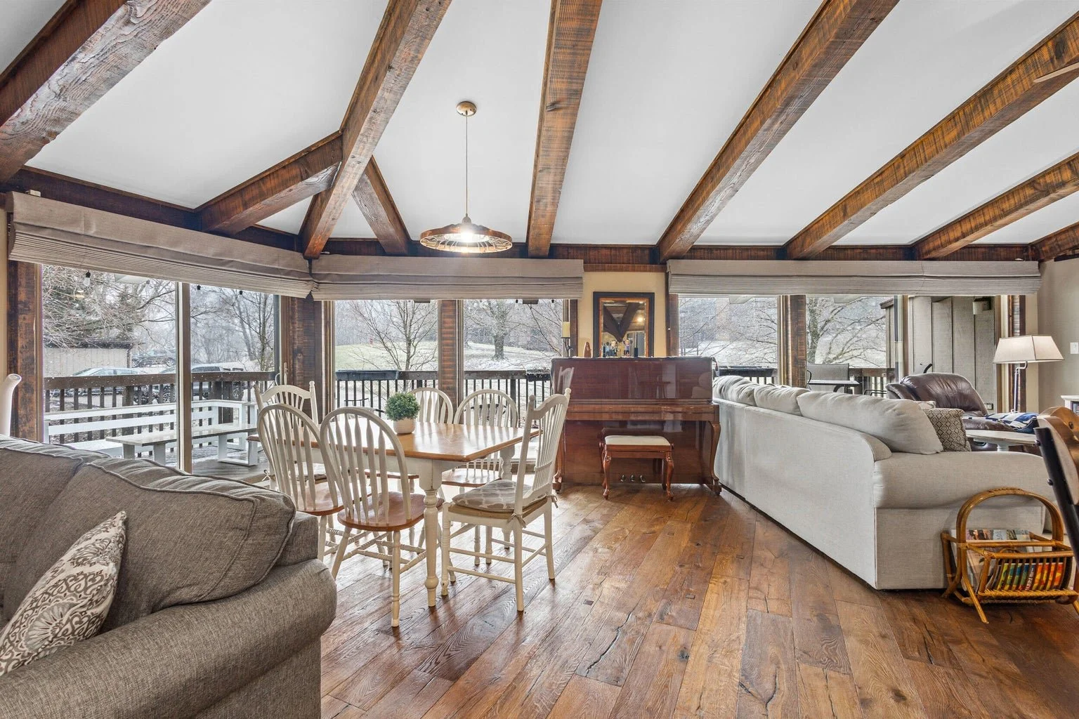 Living room with large glass windows, wooden ceiling beams, hardwood floors, white sofa, beige armchair, dining table with six chairs, piano, and snow outside.