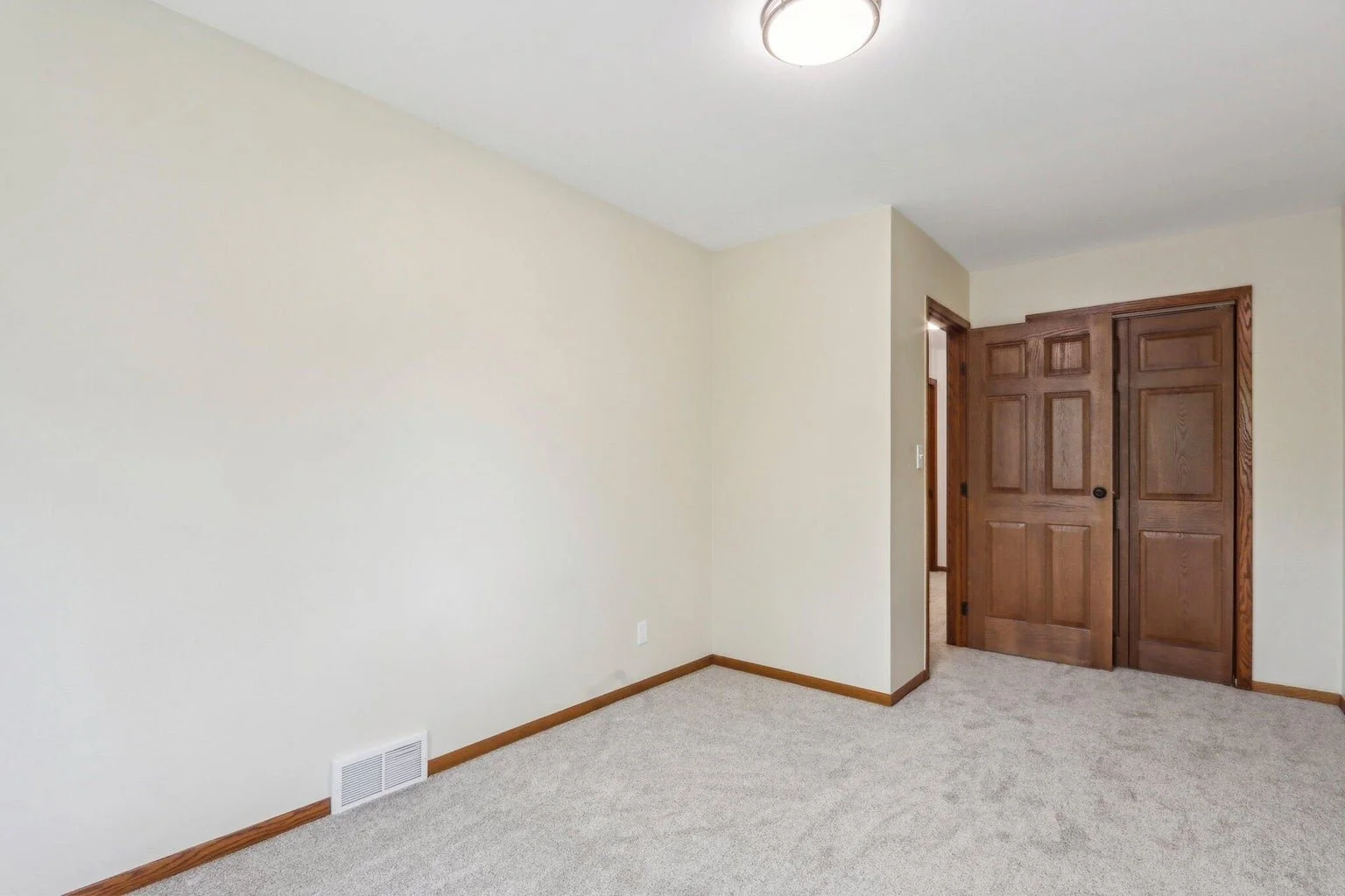 Empty room with beige carpet, white walls, wooden trim, ceiling light fixture, and brown wooden sliding door.