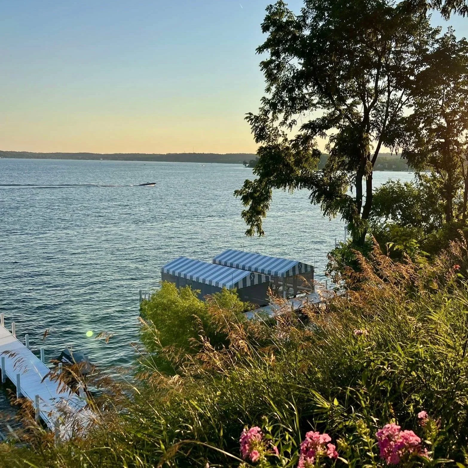 Scenic lakeside view during sunset with trees, a boat on the water, and striped canopies over a dock and seating area.
