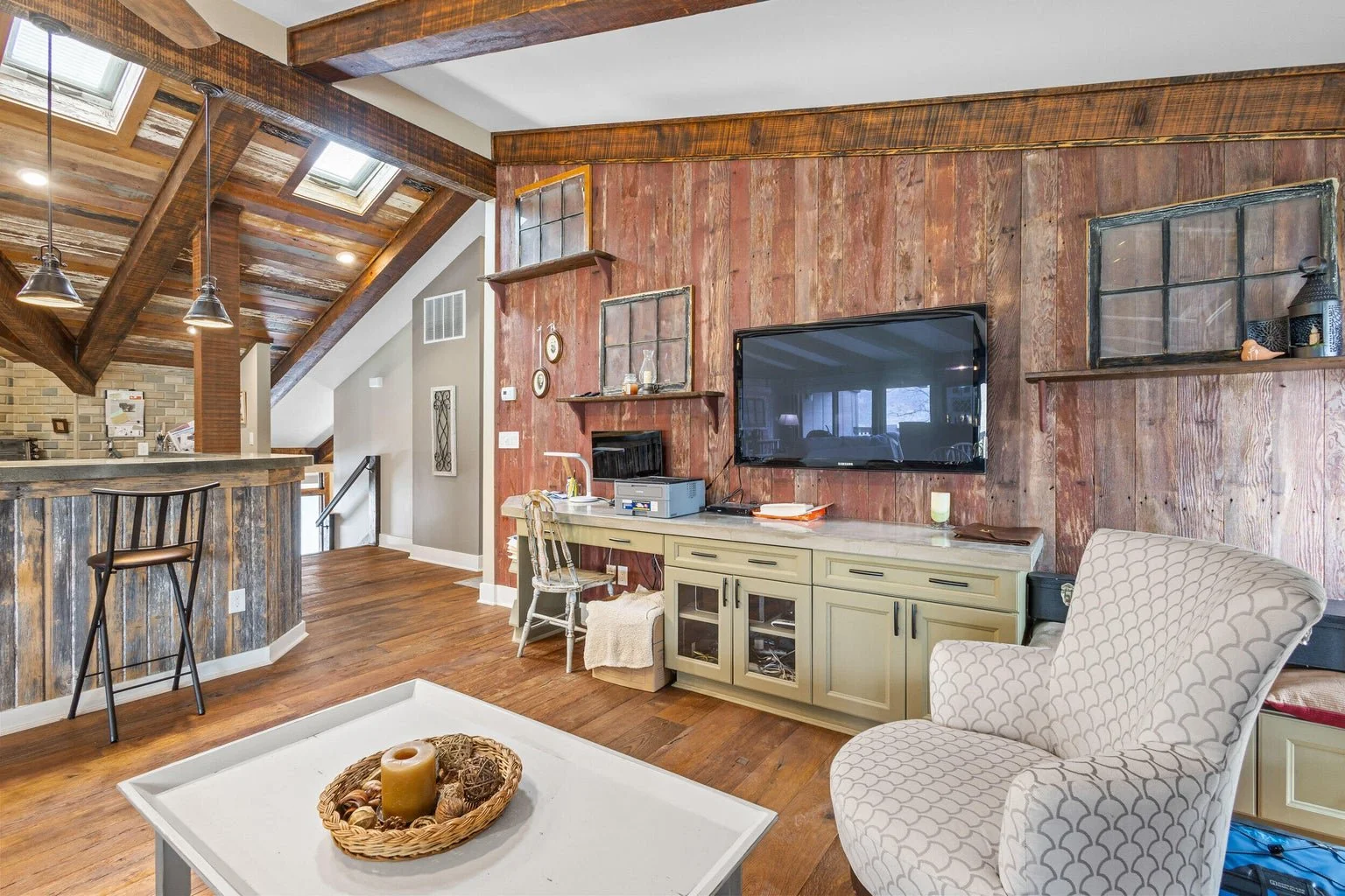 Living room with wood-paneled wall, mounted flat-screen TV, cream cabinet, patterned armchair, white coffee table with candles, and rustic decor with shelves.