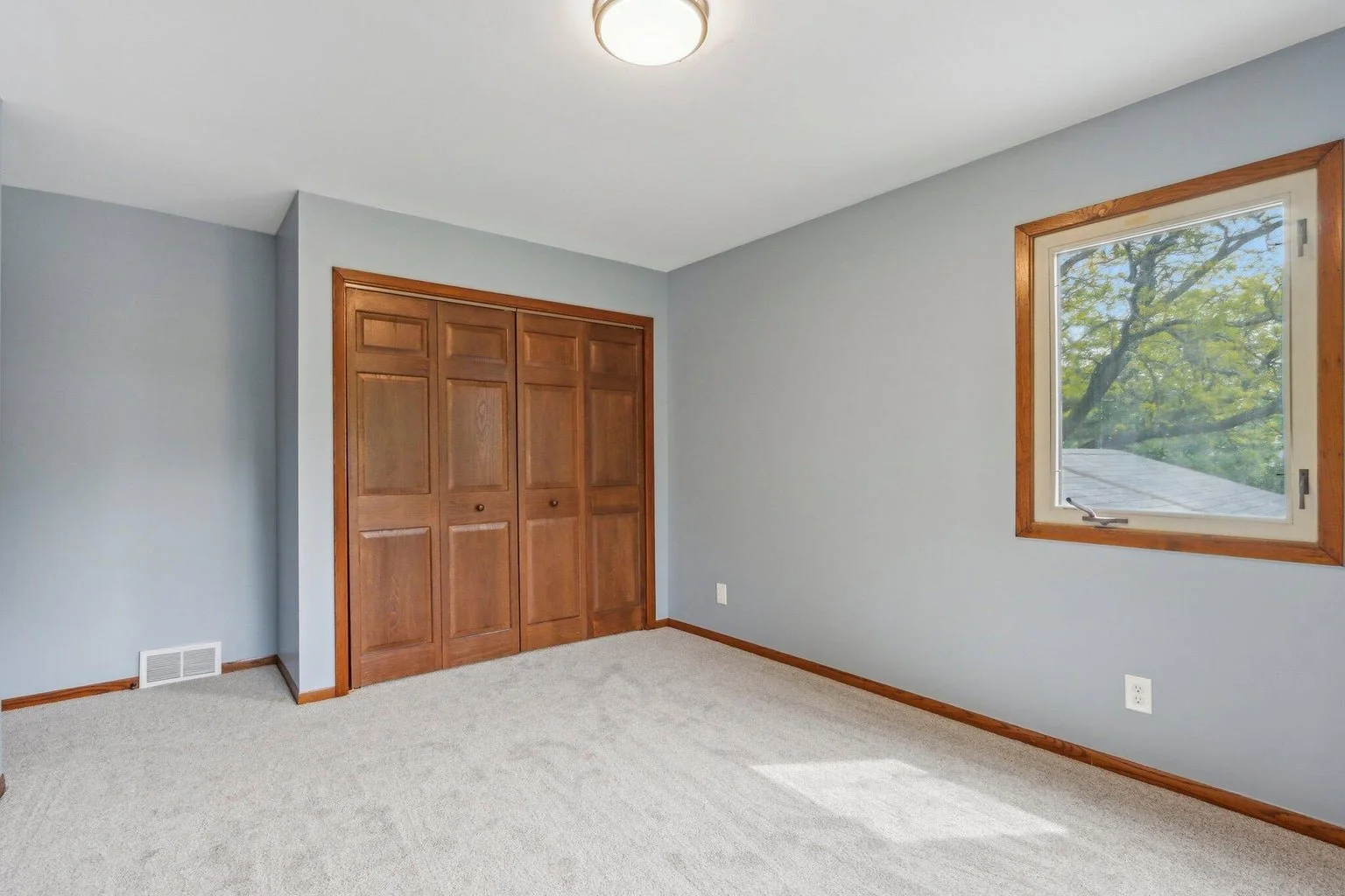 Empty bedroom with light gray walls, wooden trim, a closed set of closet doors, a window with a view of trees, beige carpet, and a ceiling light fixture.